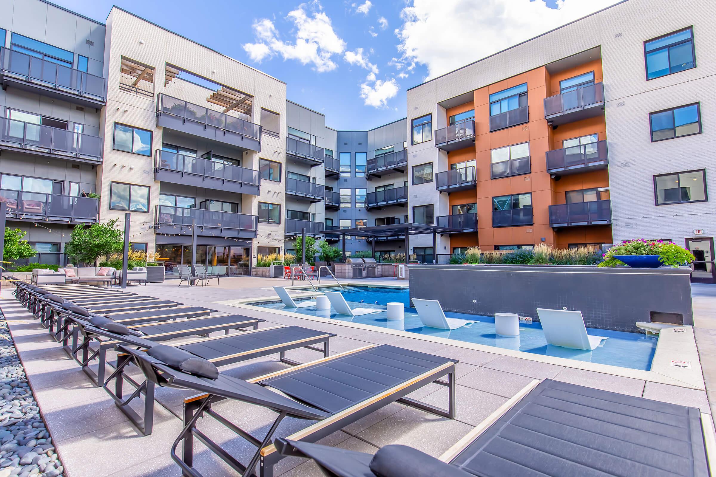 A modern apartment complex featuring a central courtyard with a swimming pool. Lounge chairs are arranged around the pool area, and balconies are visible on the surrounding buildings. Green plants and trees add a touch of nature, while clear blue skies complement the contemporary architecture.