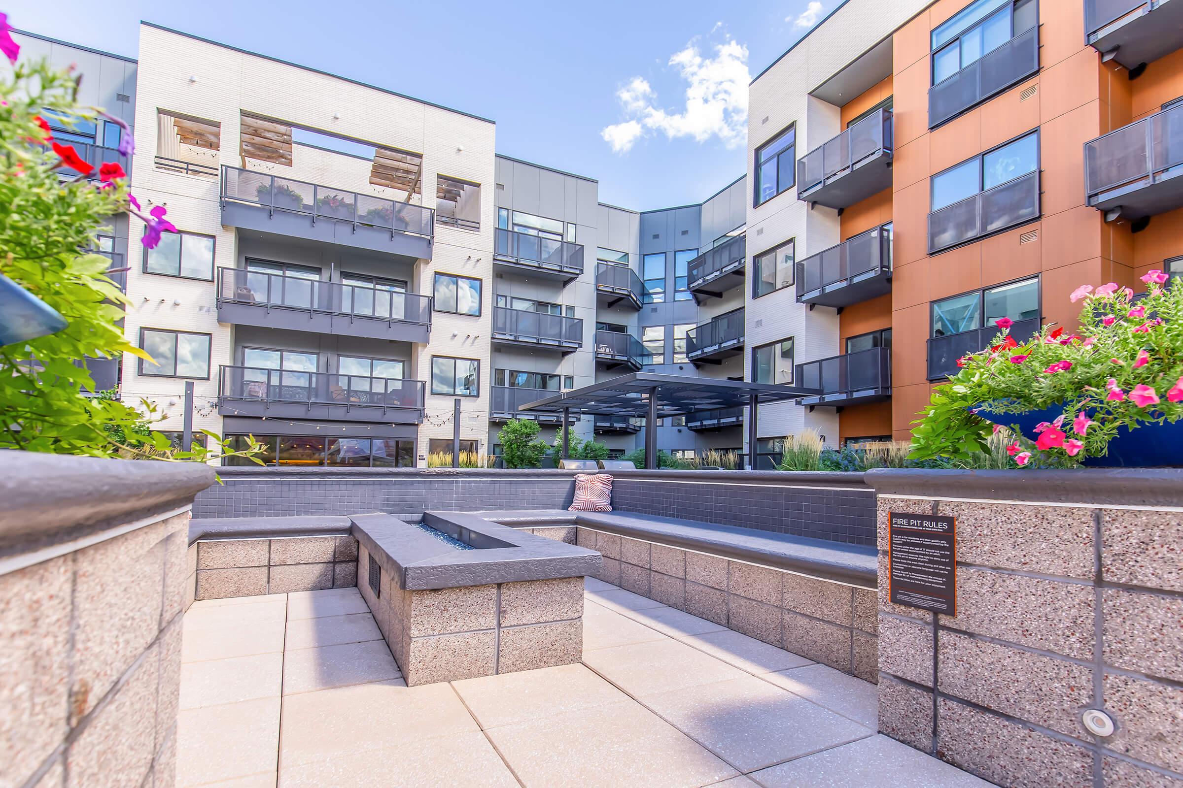 A modern apartment complex featuring outdoor seating and lush landscaping. The surrounding area includes colorful flower pots, spacious balconies, and multiple levels of the building, with an inviting communal space in the foreground, perfect for relaxation. Bright blue sky visible above.