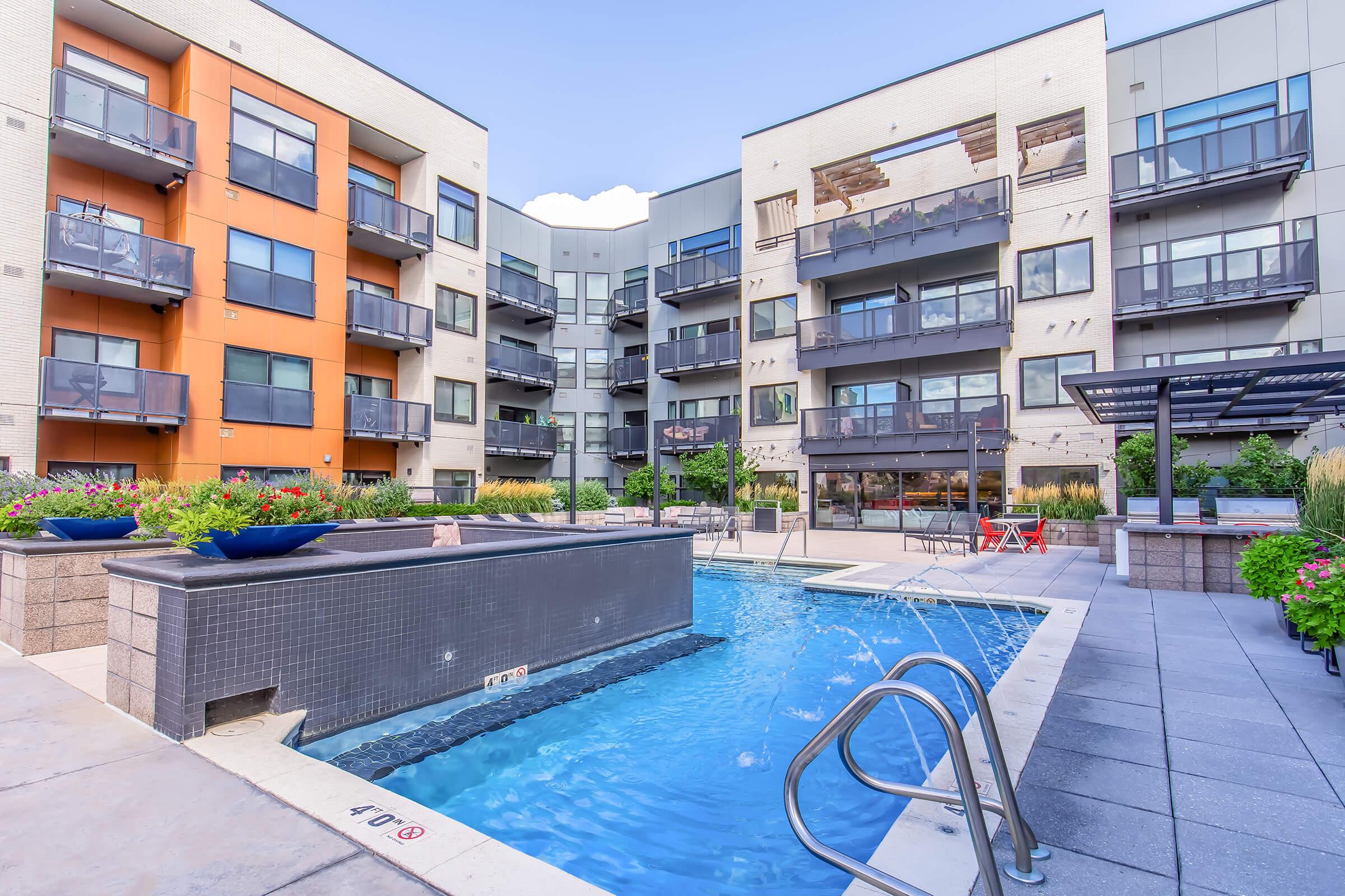 Modern apartment complex courtyard featuring a swimming pool with a waterfall, surrounded by well-maintained gardens and seating areas. The buildings showcase a mix of brick and contemporary grey facades, with balconies overlooking the pool area. Ideal setting for relaxation and social gatherings.