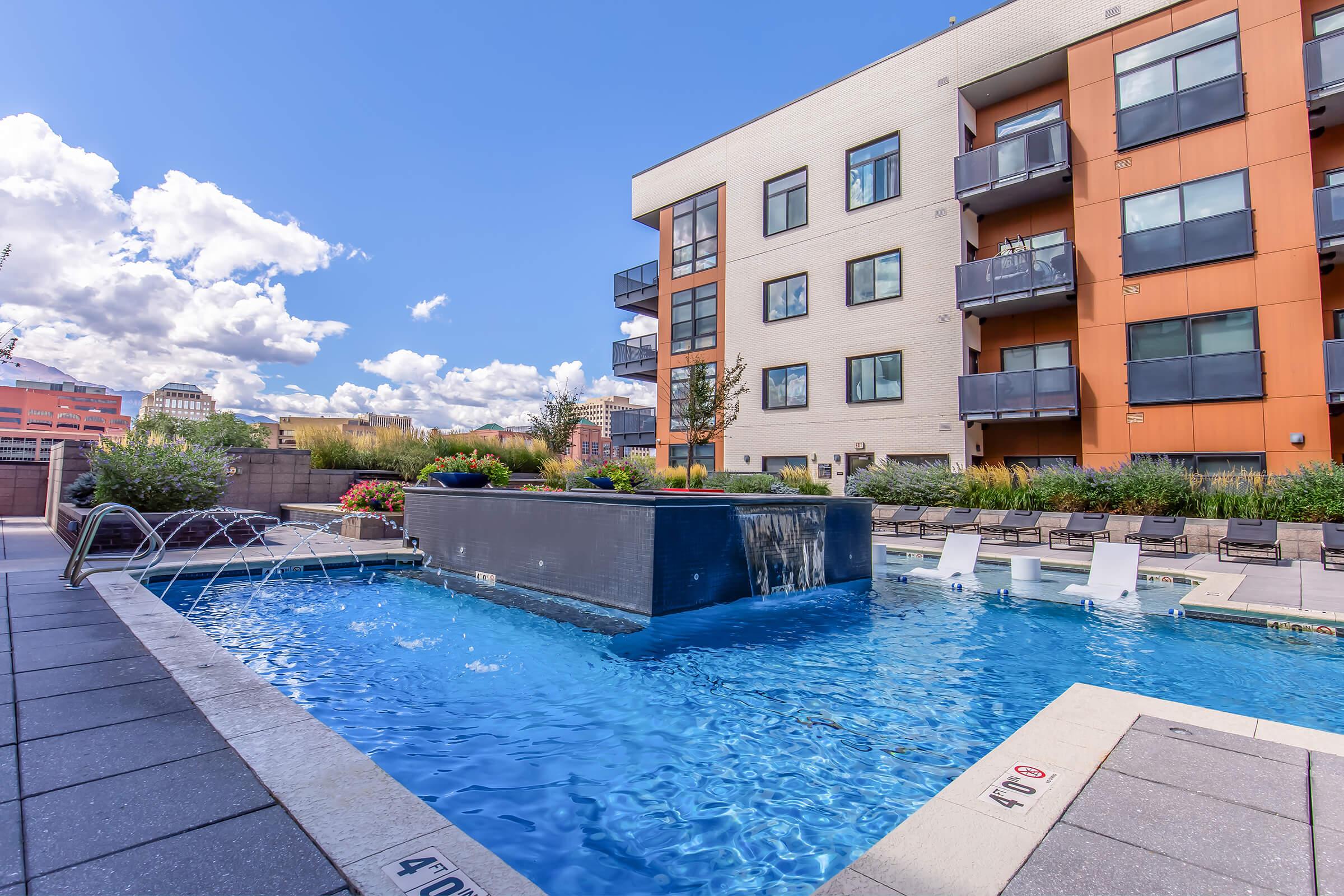 A modern apartment complex featuring a swimming pool with a waterfall. The pool area is surrounded by lounge chairs and landscaped greenery. The sky is partly cloudy, adding to the inviting atmosphere of the outdoor space.