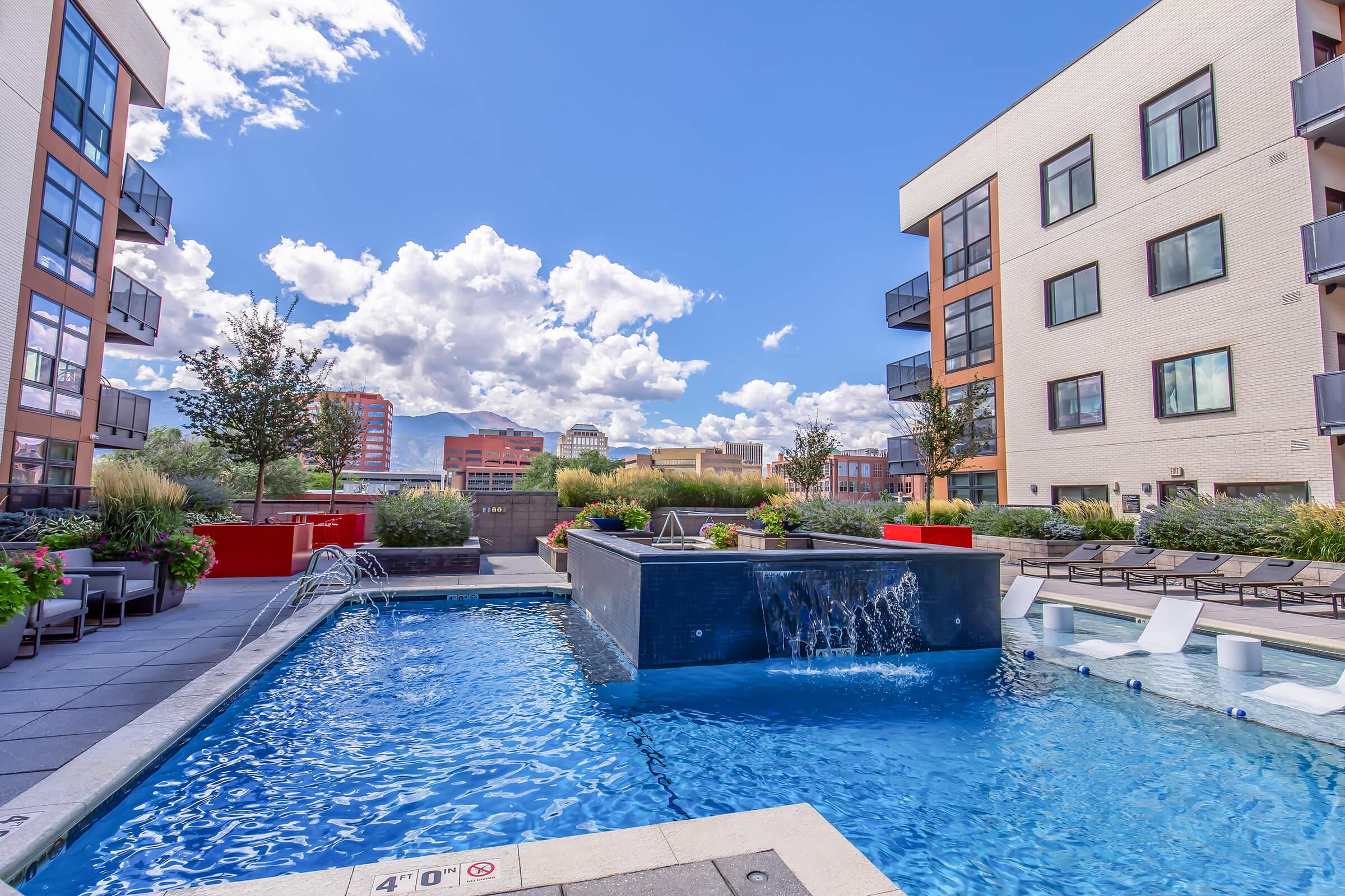 A modern outdoor pool area featuring a large blue swimming pool with a waterfall, surrounded by lounge chairs and greenery. Nearby, there are stylish apartment buildings with large windows and balconies, set against a vibrant blue sky with fluffy white clouds.
