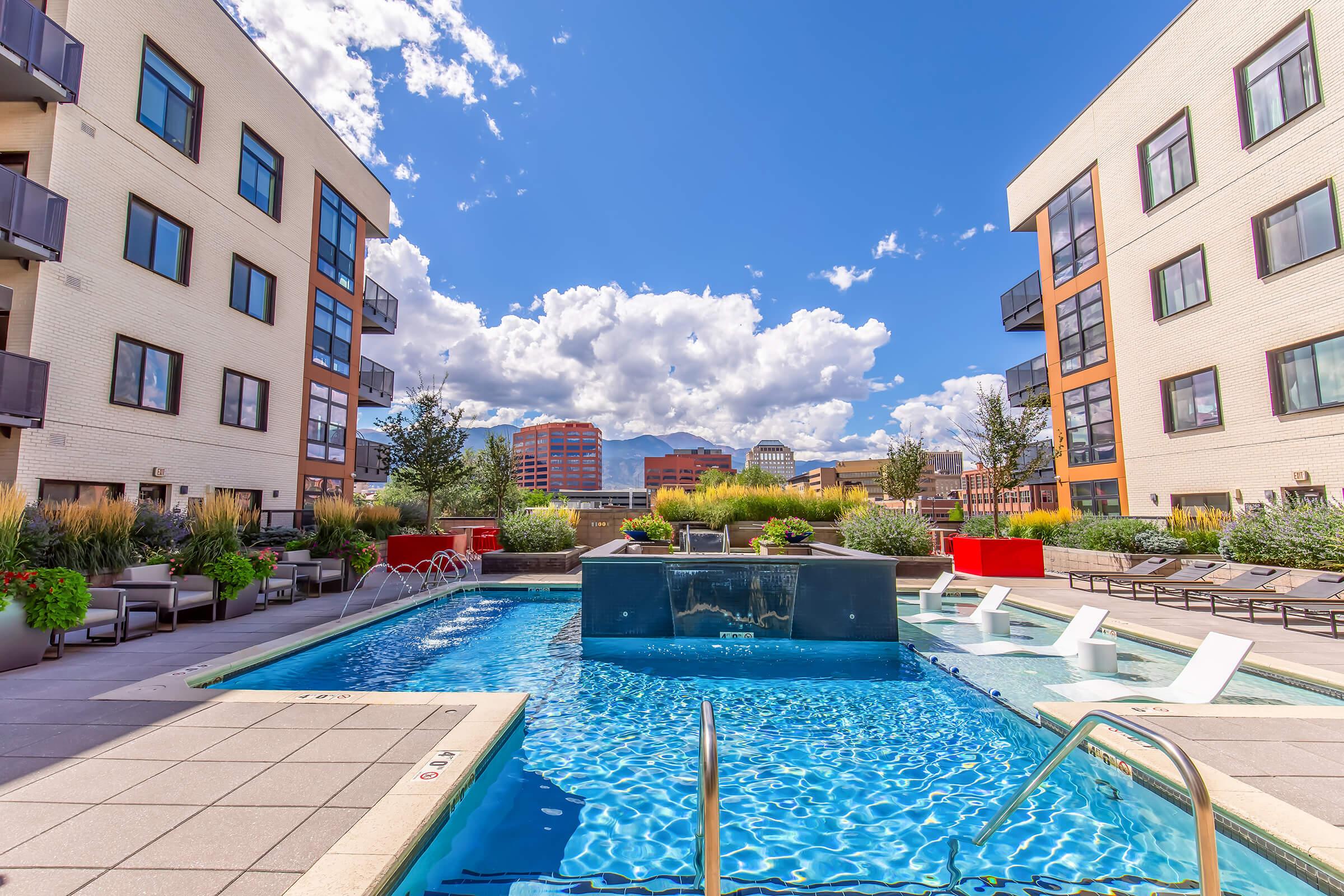 A serene apartment complex courtyard featuring a sparkling blue swimming pool, surrounded by green landscaping, lounge chairs, and trees. In the background, modern buildings and a clear blue sky with fluffy white clouds create a vibrant atmosphere.