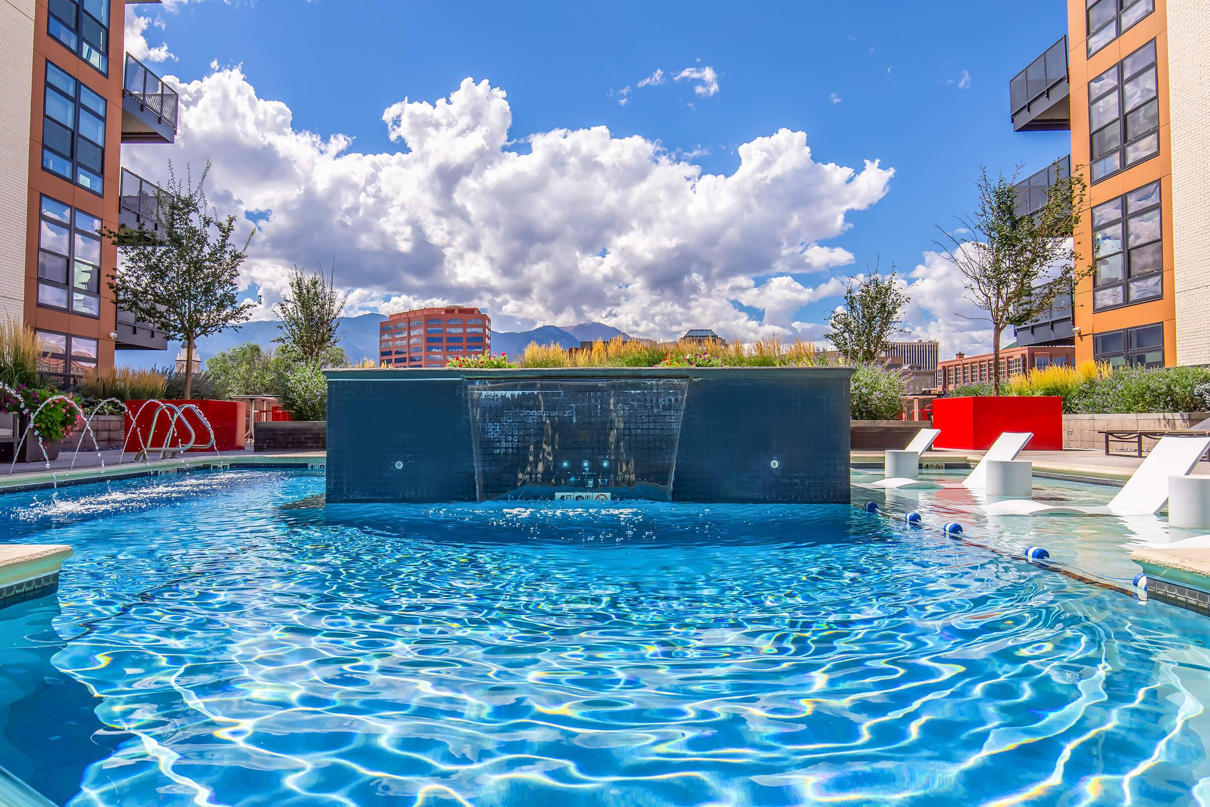 A vibrant swimming pool surrounded by modern apartment buildings, featuring a waterfall wall, lounge chairs, and lush greenery. The sky is bright blue with fluffy white clouds and distant mountains in the background, creating a picturesque and inviting atmosphere.