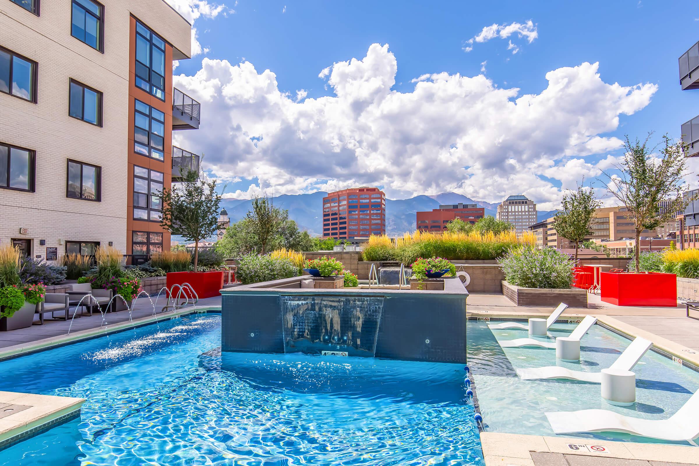 A modern outdoor swimming pool featuring a water fountain, surrounded by lush greenery and contemporary landscaping. The view includes distant mountains under a bright blue sky with fluffy white clouds, complemented by stylish apartment buildings in the background.