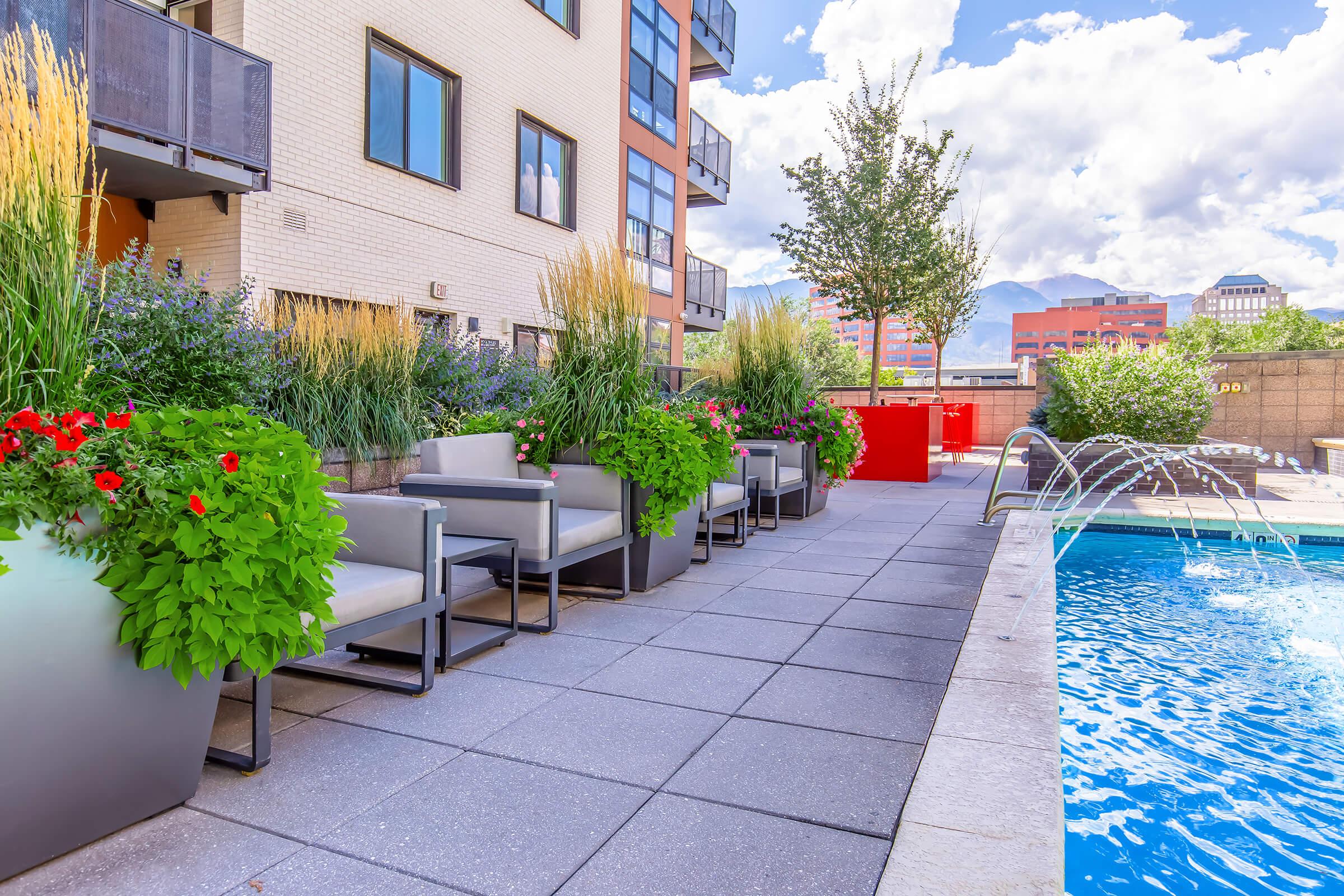 A modern outdoor area featuring a swimming pool surrounded by lush planters with vibrant flowers. Comfortable seating is arranged next to the pool, and a fountain adds a dynamic element. The scene is set against a backdrop of contemporary apartment buildings and a clear blue sky.