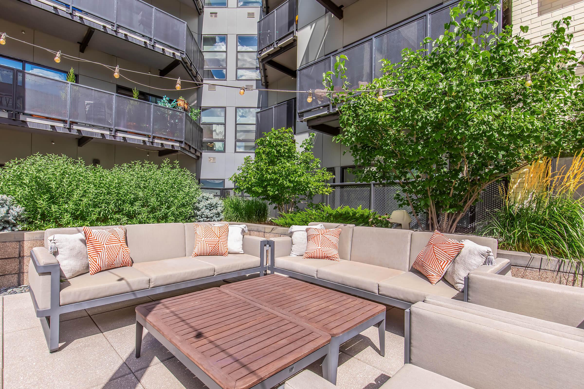 Outdoor lounge area featuring a stylish sectional sofa with orange and white patterned cushions, a wooden coffee table, and lush greenery in a modern apartment complex setting. String lights hang above, adding a cozy ambiance to the space.