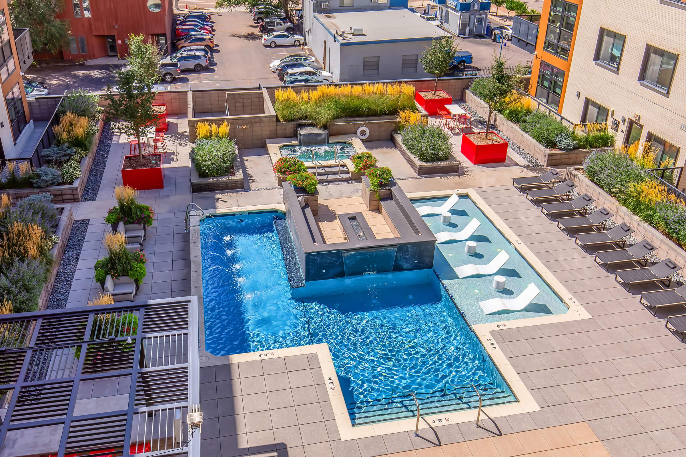Aerial view of a modern pool area featuring a rectangular swimming pool with lounge chairs nearby, hot tubs, and landscaped greenery. Surrounding the pool are red planters and stylish seating areas, all situated within a contemporary apartment complex setting.