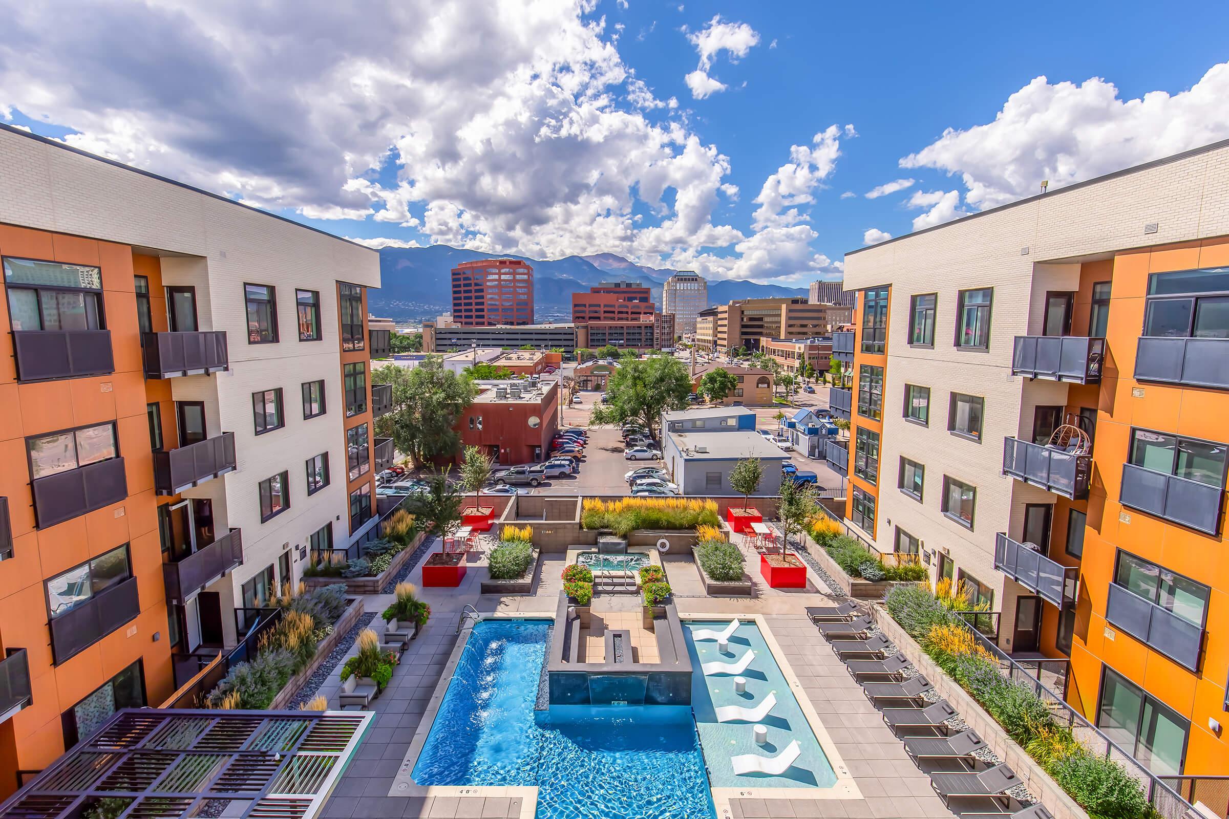 View of a modern apartment complex with orange and white buildings surrounding a swimming pool. In the background, a city skyline is visible under a blue sky with fluffy clouds. The pool area features lounge chairs and landscaped gardens.