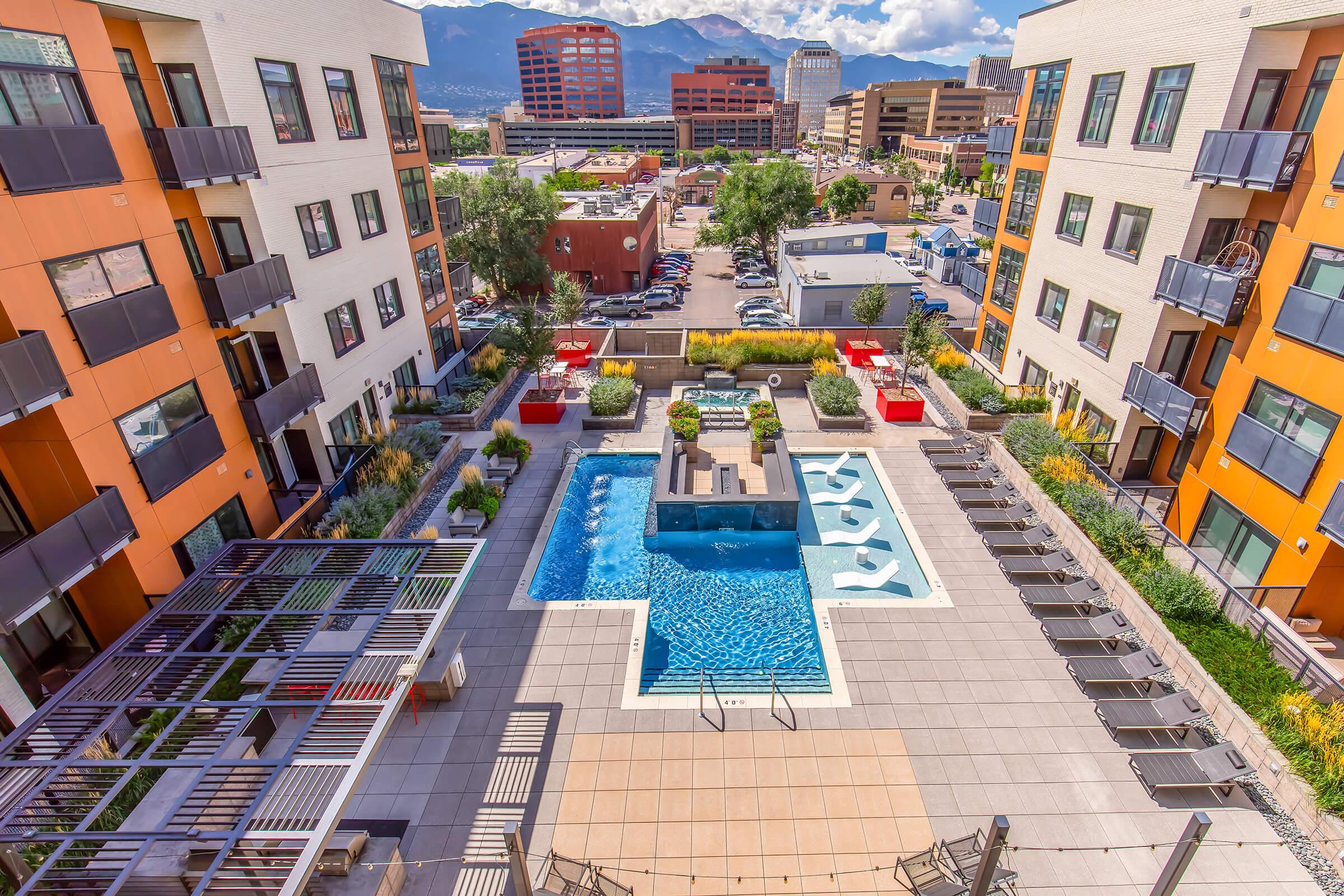 Aerial view of an apartment complex courtyard featuring a swimming pool with a modern design, surrounded by lounge chairs and red seating areas. Lush greenery and landscaping enhance the outdoor space, with cityscape and mountains visible in the background. Bright, sunny day with blue skies.