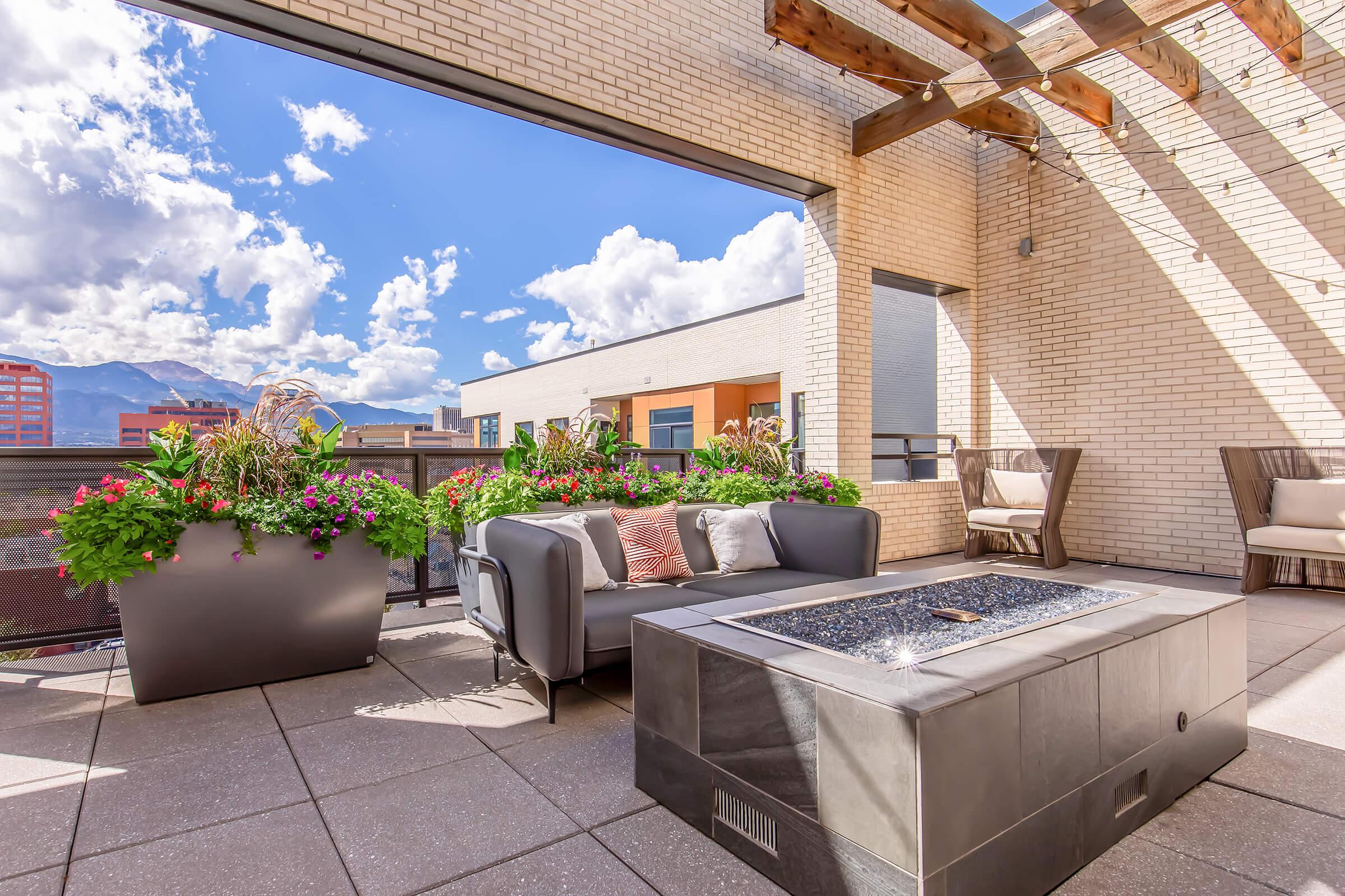 A modern rooftop patio featuring a stylish sofa with decorative pillows, a fire pit with a glass top, vibrant flower planters, and comfortable seating. The space is surrounded by views of buildings and a blue sky with fluffy white clouds, creating a relaxing outdoor atmosphere.