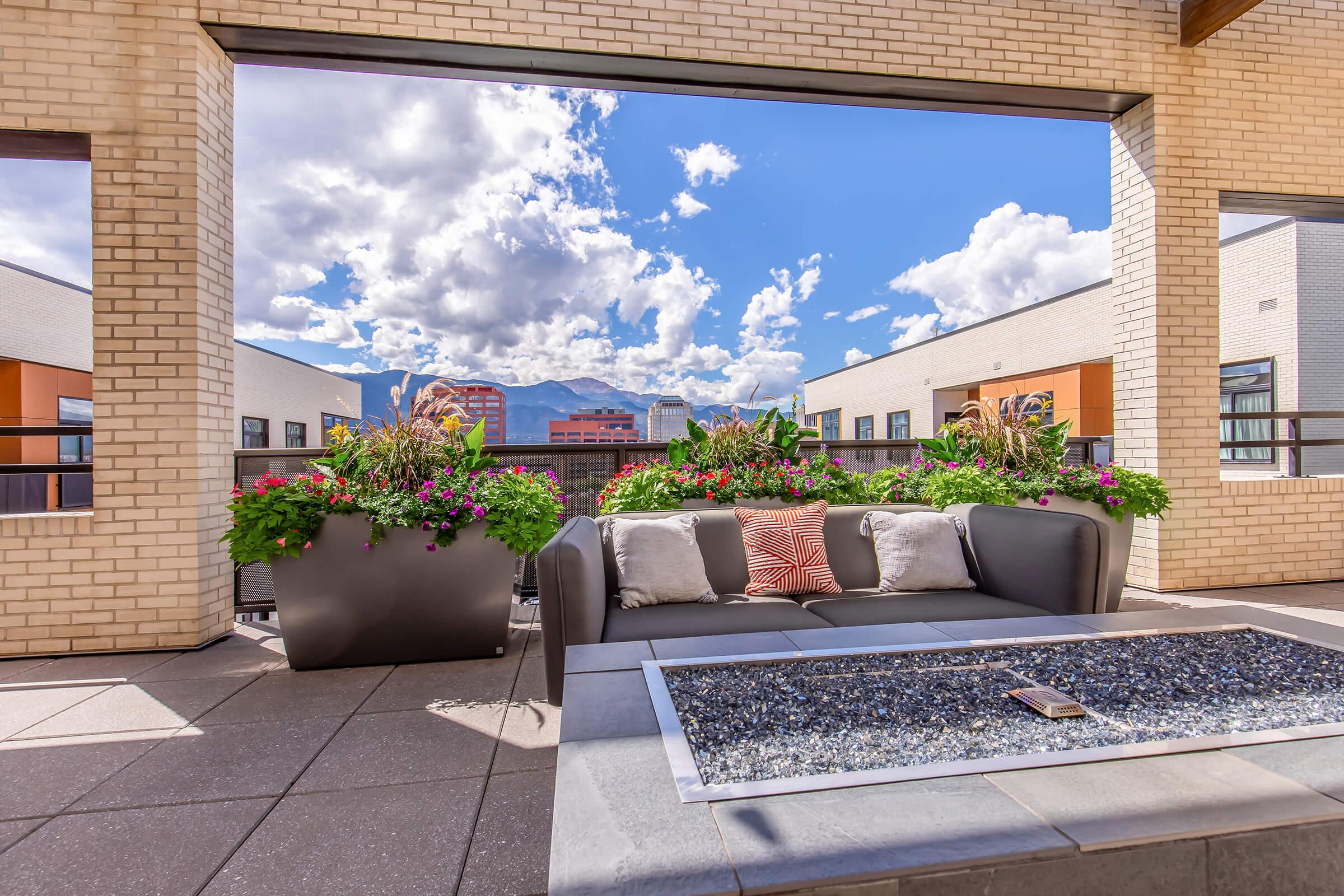 A stylish outdoor lounge area featuring a modern sofa with decorative cushions, set on a rooftop patio. The space is adorned with vibrant flower pots and offers a panoramic view of the sky filled with fluffy clouds and surrounding buildings, creating a serene and inviting atmosphere.