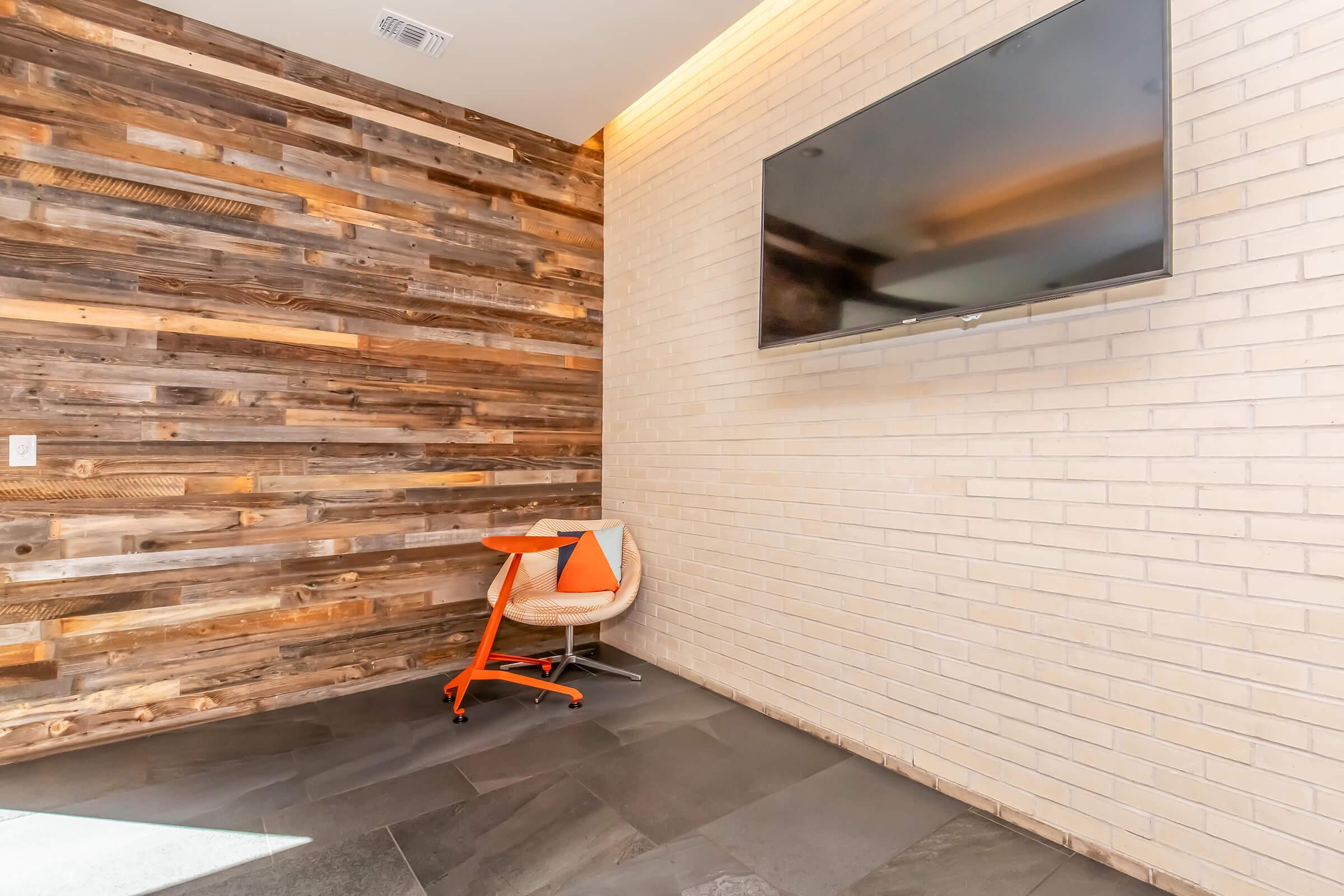 A modern interior space featuring a wooden accent wall, a flat-screen TV mounted on a light-colored brick wall, and a small round table with a chair. The floor is covered with dark tiles, creating a stylish and contemporary atmosphere.