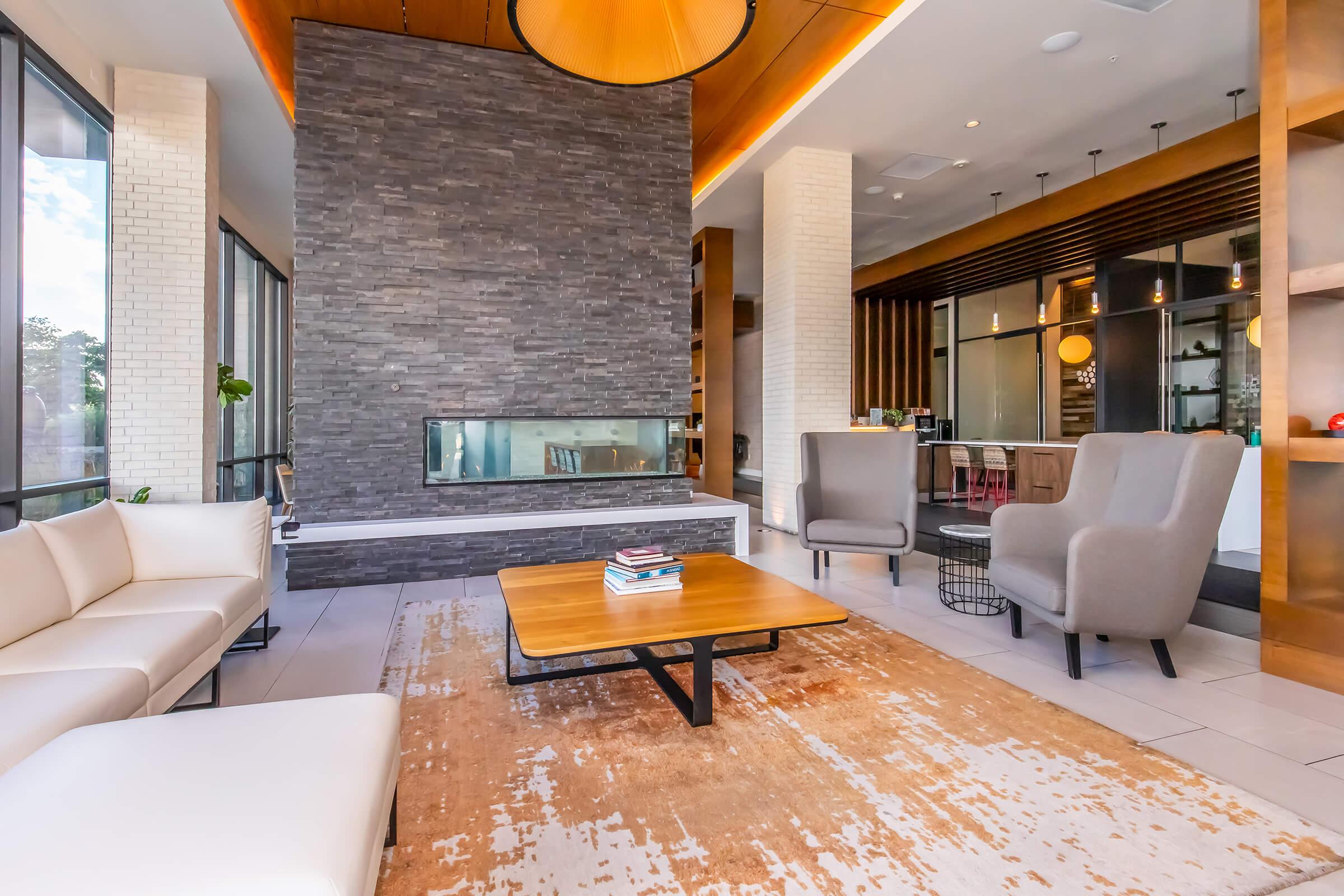 Cozy modern lounge area featuring a stylish fireplace, large windows allowing natural light, a beige area rug, a wooden coffee table, and two gray armchairs. In the background, there's a hint of a contemporary kitchen and dining space, creating an inviting atmosphere.