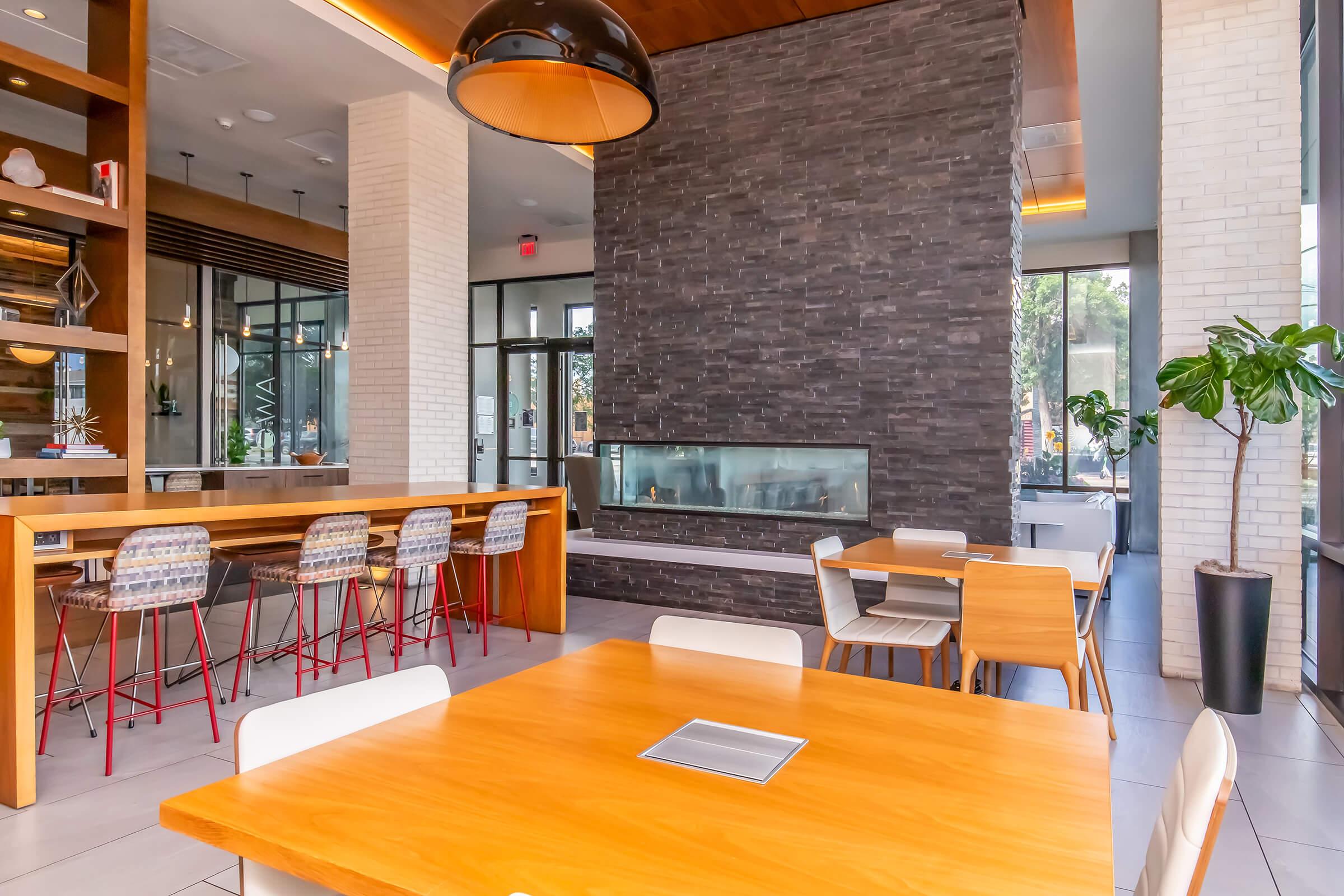 Modern dining area featuring wooden tables and chairs, a sleek bar with high stools, and a stylish stone fireplace. Large windows allow natural light to fill the space, enhanced by contemporary decor and greenery. Ideal for casual dining or social gatherings.