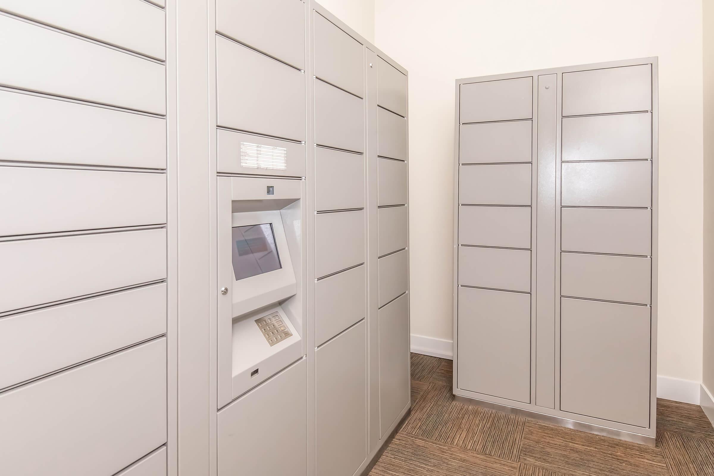 A modern package delivery room featuring multiple gray lockers for secure storage. One locker unit includes an electronic keypad or screen for accessing packages. The floor is brown with a textured design, and the walls are painted in a light color, creating a clean and organized environment.