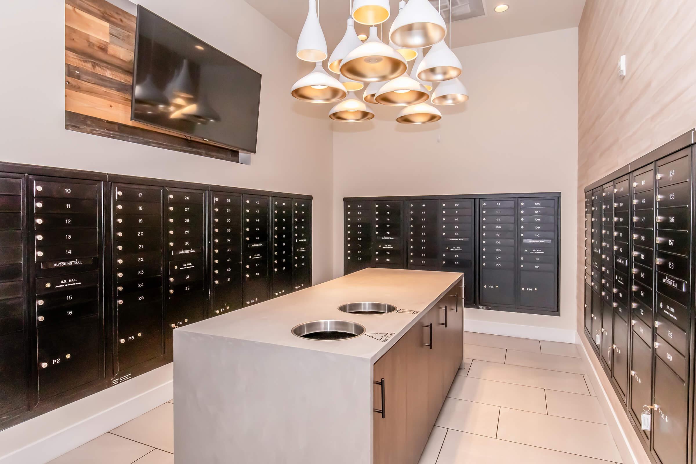 A modern package delivery room featuring black mail lockers lining the walls, a centrally located concrete table with round openings, and stylish pendant lighting above. A flat-screen TV is mounted on the back wall, creating a contemporary and functional space for receiving packages.
