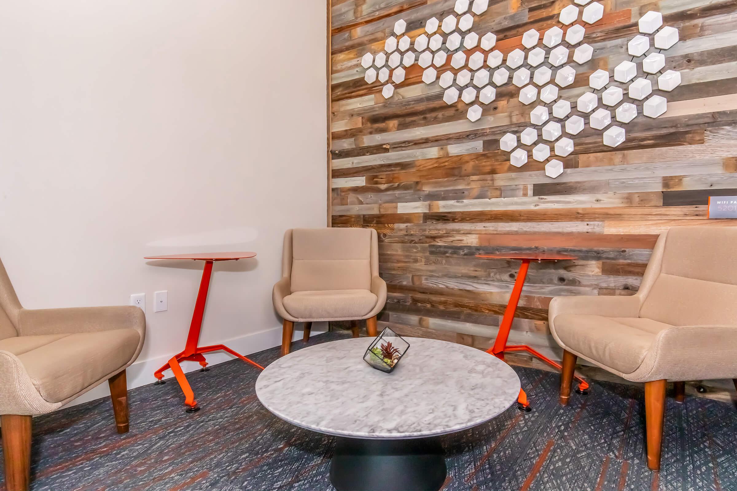 A modern seating area featuring two beige chairs and a round marble table on a patterned carpet. The background consists of a rustic wooden wall with hexagonal white decor elements. The space is designed for relaxation or informal gatherings.