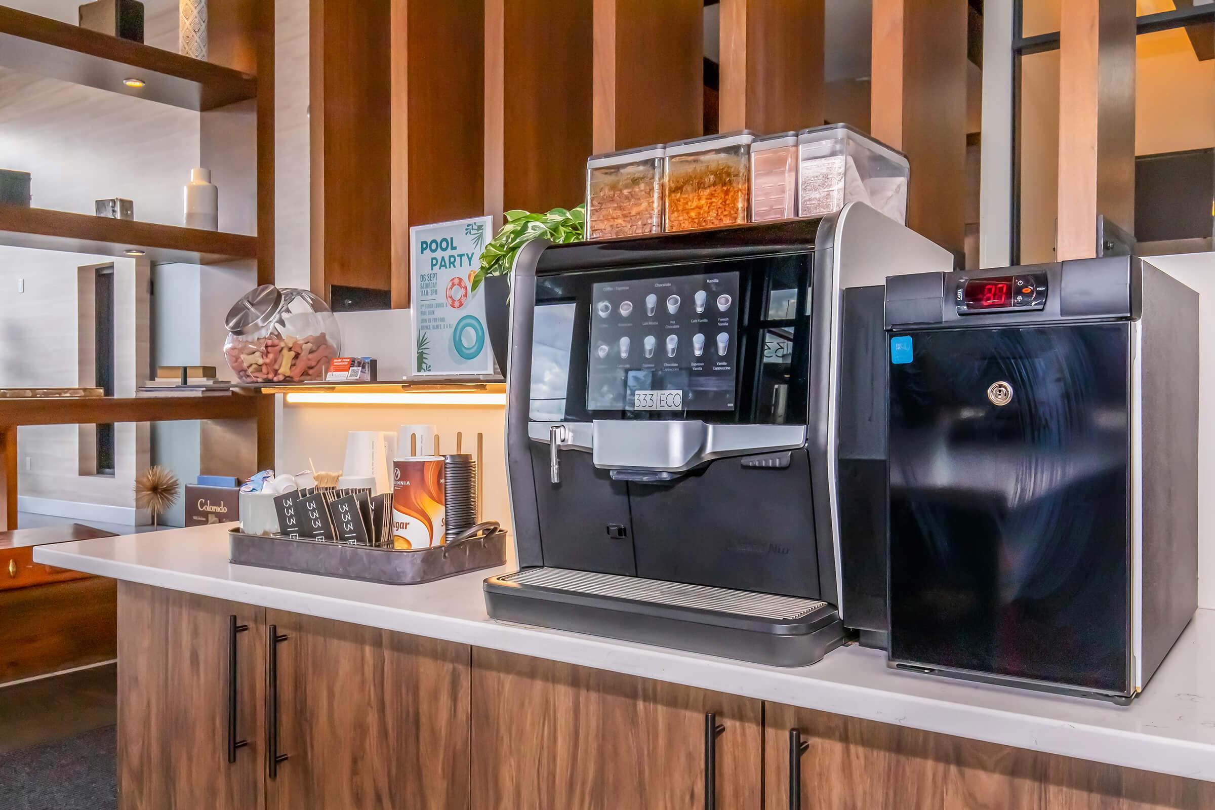 A modern coffee machine with a digital display sits on a counter next to a black refrigerator. Above the coffee machine are clear containers filled with various ingredients. The setting is well-lit with wooden paneling and a few decorative items in the background. A sign for a "Pool Party" is visible.