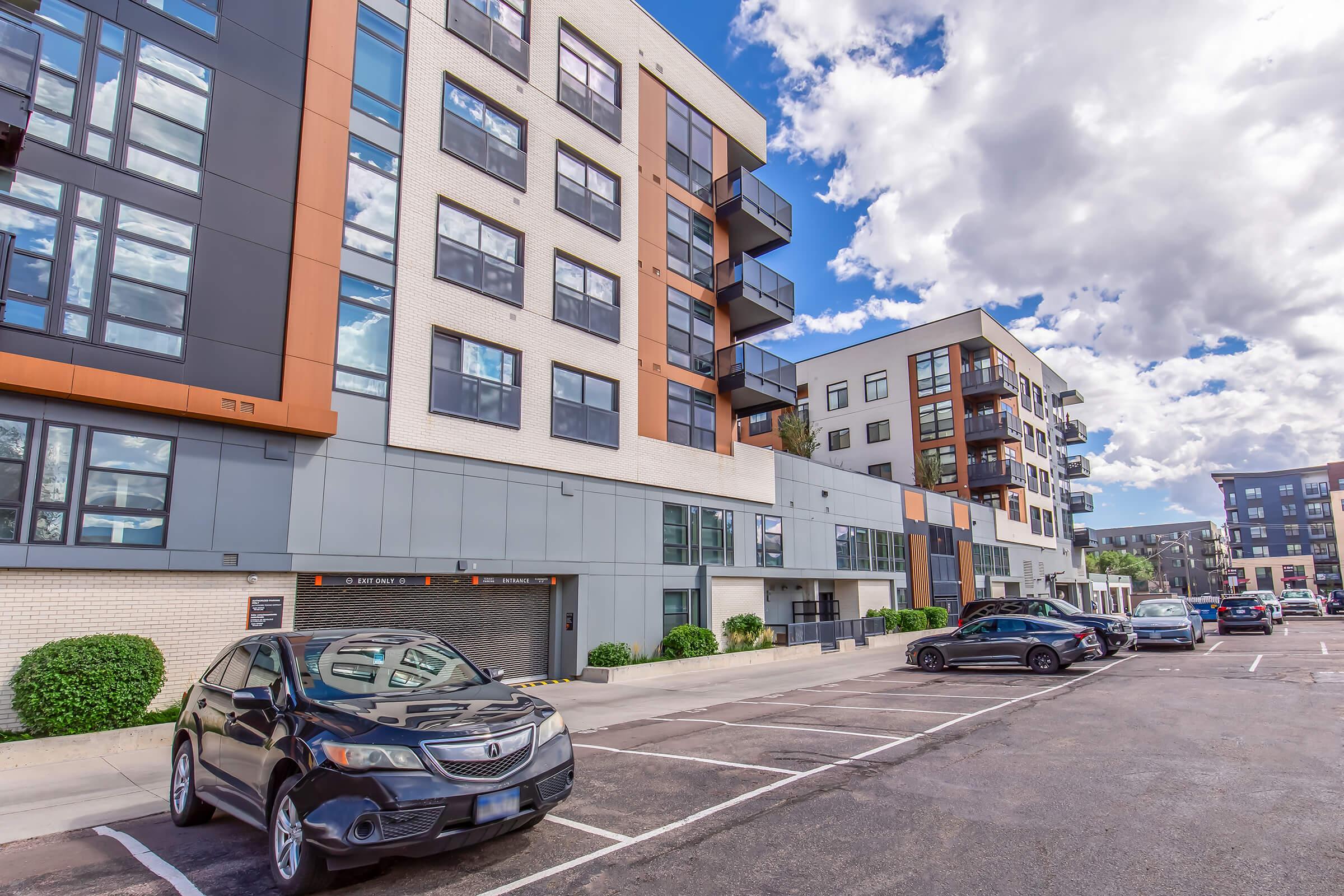 A modern apartment complex featuring contemporary architecture with large windows and a mix of brick and metal facades. The scene includes a parking lot with several cars parked, surrounded by neatly trimmed bushes and a partly cloudy blue sky.