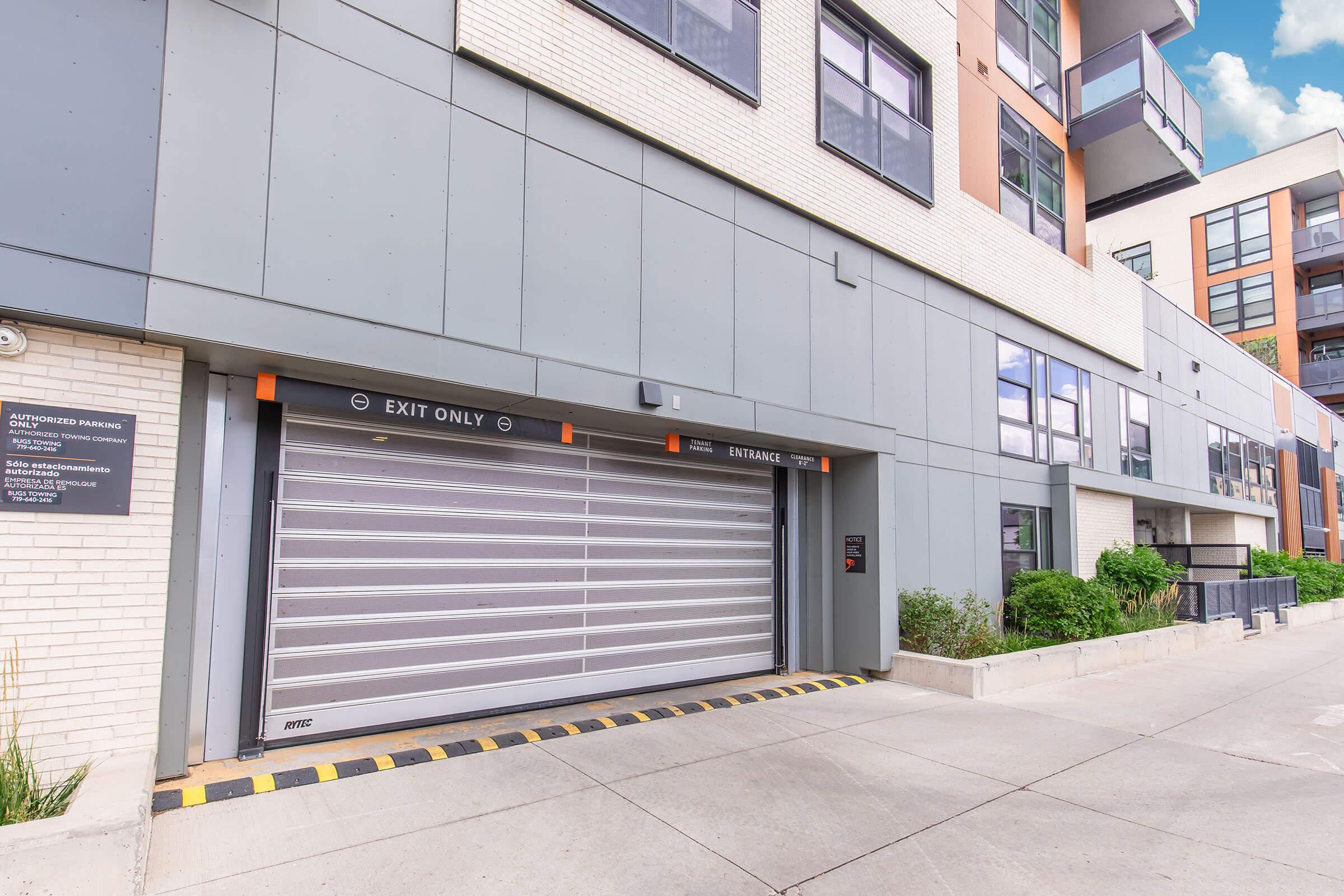 A modern building entrance featuring a secure, rolled-up garage door labeled "EXIT ONLY." The exterior is a mix of light-colored brick and gray panels, with large windows reflecting the sky. Green plants are visible near the entrance, and a sign is mounted on the wall nearby, indicating parking regulations.