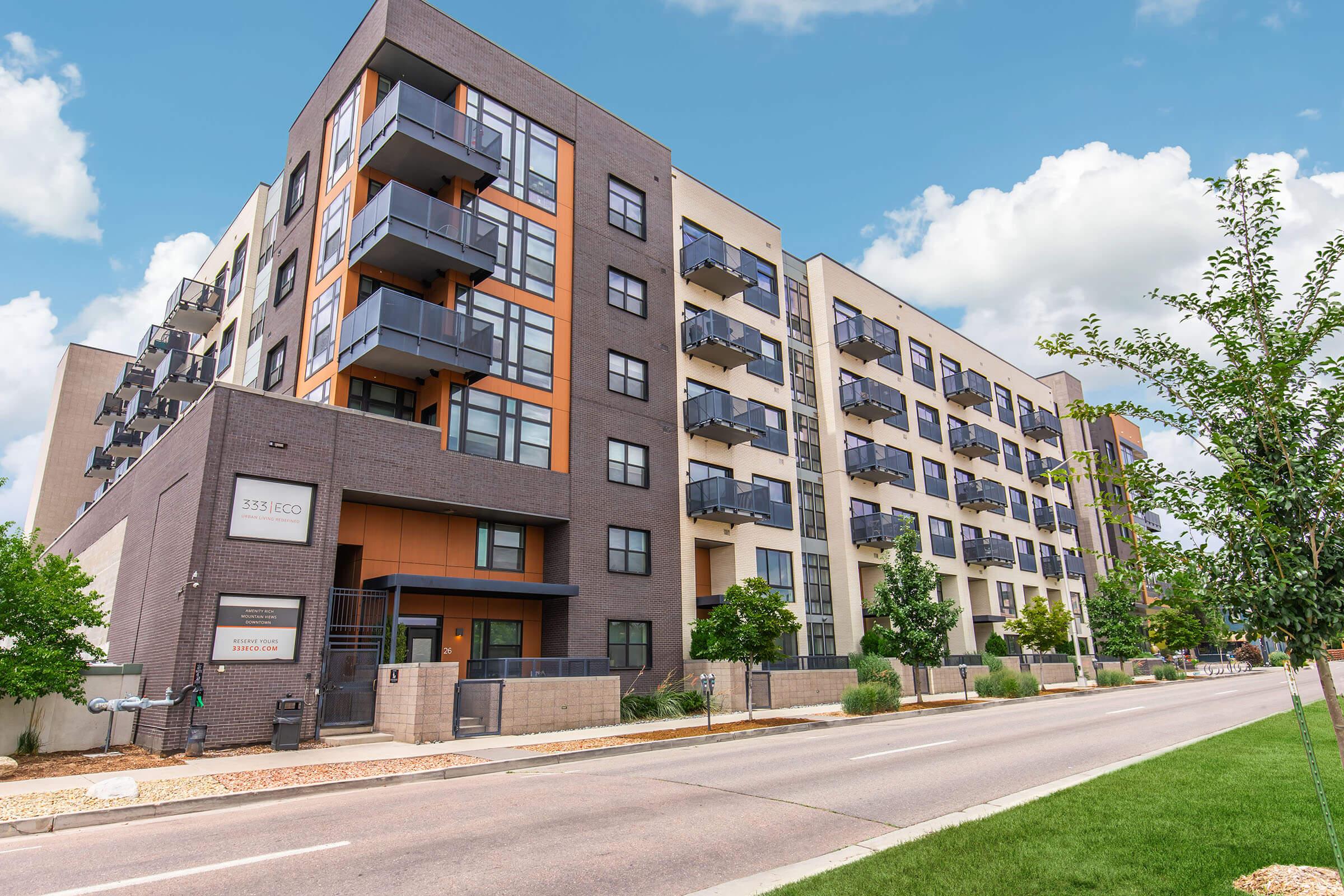 A modern apartment building with a mix of brick and stucco exteriors, featuring multiple balconies and large windows. The building is set along a street lined with greenery, and a clear blue sky with scattered clouds is visible in the background. The entrance is marked with a sign.