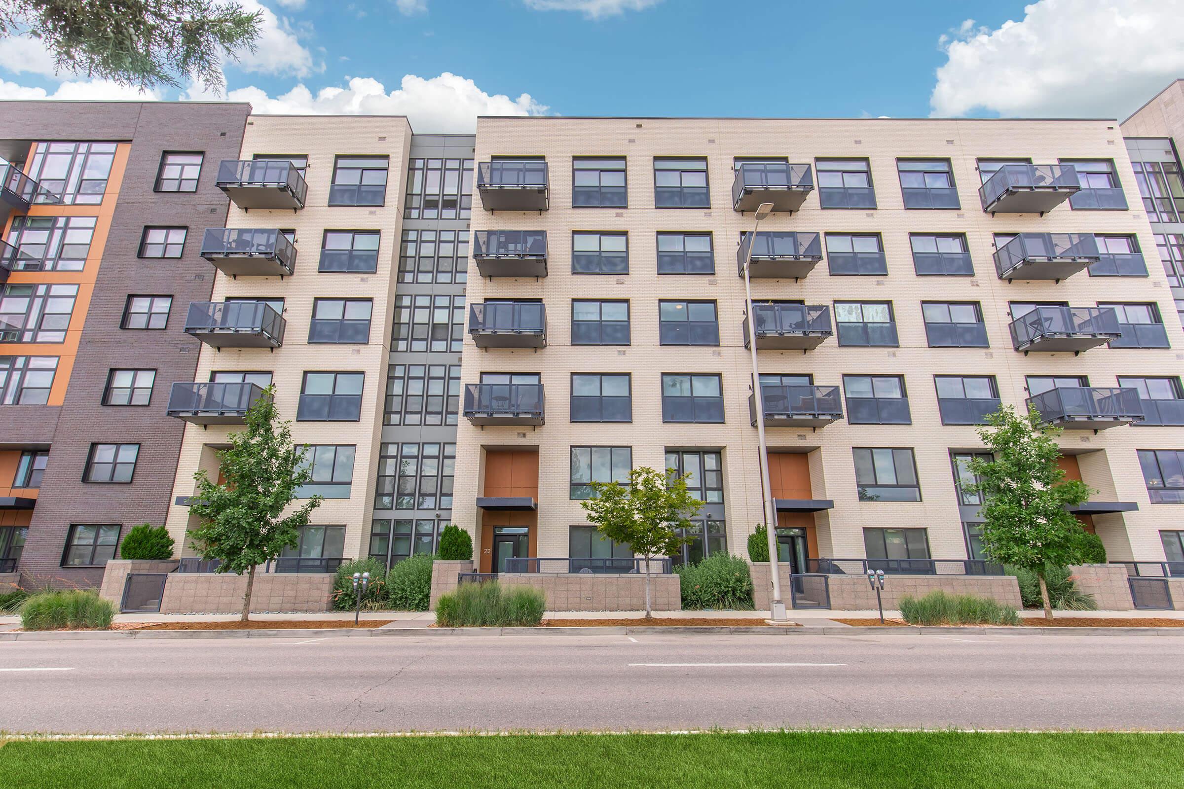 Modern apartment building with multiple stories and balconies, featuring large windows. The facade includes a mix of light and dark materials, complemented by trees and landscaping along the sidewalk. The sky is blue with a few clouds, and a street runs in front of the building.