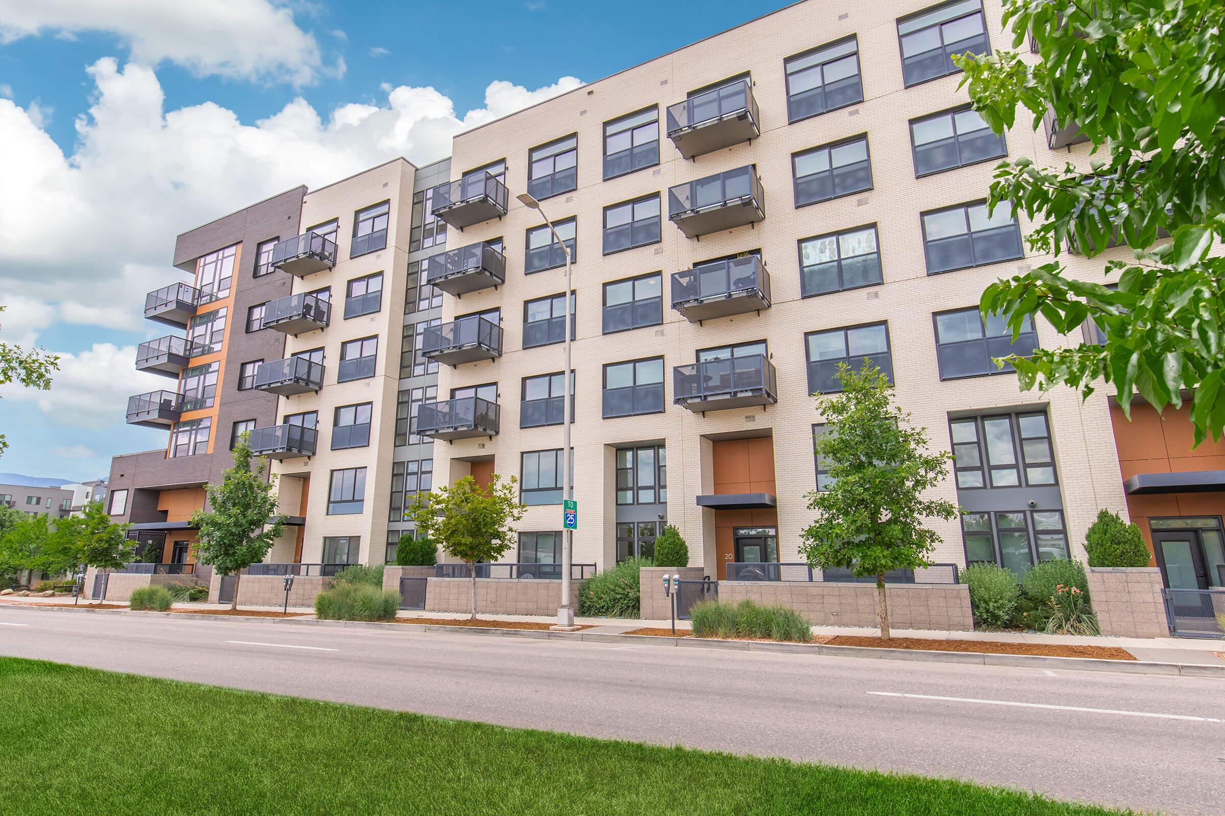 Modern residential building featuring a mix of brick and siding, with large windows and multiple balconies. There are landscaped shrubs along the sidewalk, and the scene is set against a partly cloudy blue sky. The building has four stories, with a clean and contemporary design.