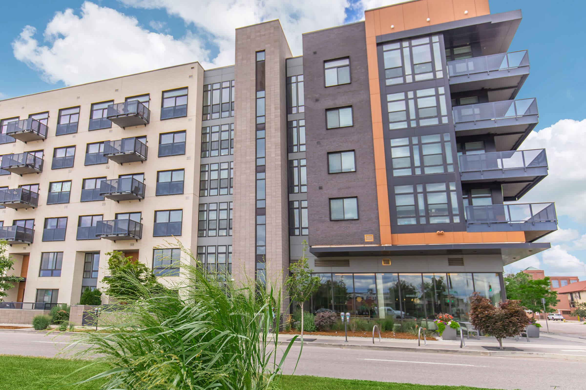 Modern apartment building with a mix of light and dark facade materials, featuring balconies, large windows, and green landscaping in the foreground. The sky is partly cloudy, adding to the vibrant atmosphere of the scene.