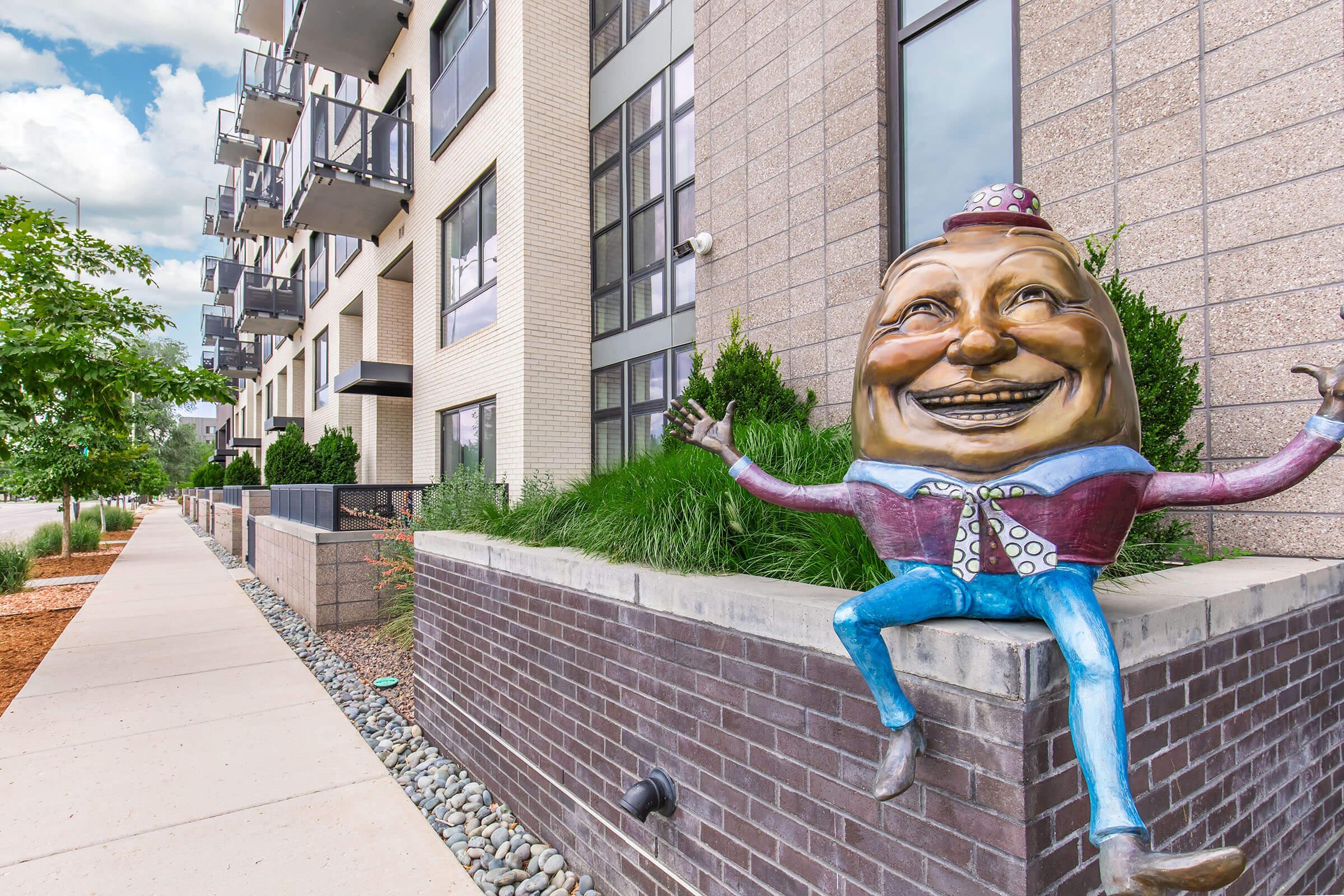 A whimsical statue of a smiling, cartoonish character in colorful attire sits on a brick wall outside a modern building. The building features large windows and balconies, surrounded by greenery and a landscaped sidewalk. The sky is partly cloudy, adding a vibrant backdrop to the scene.
