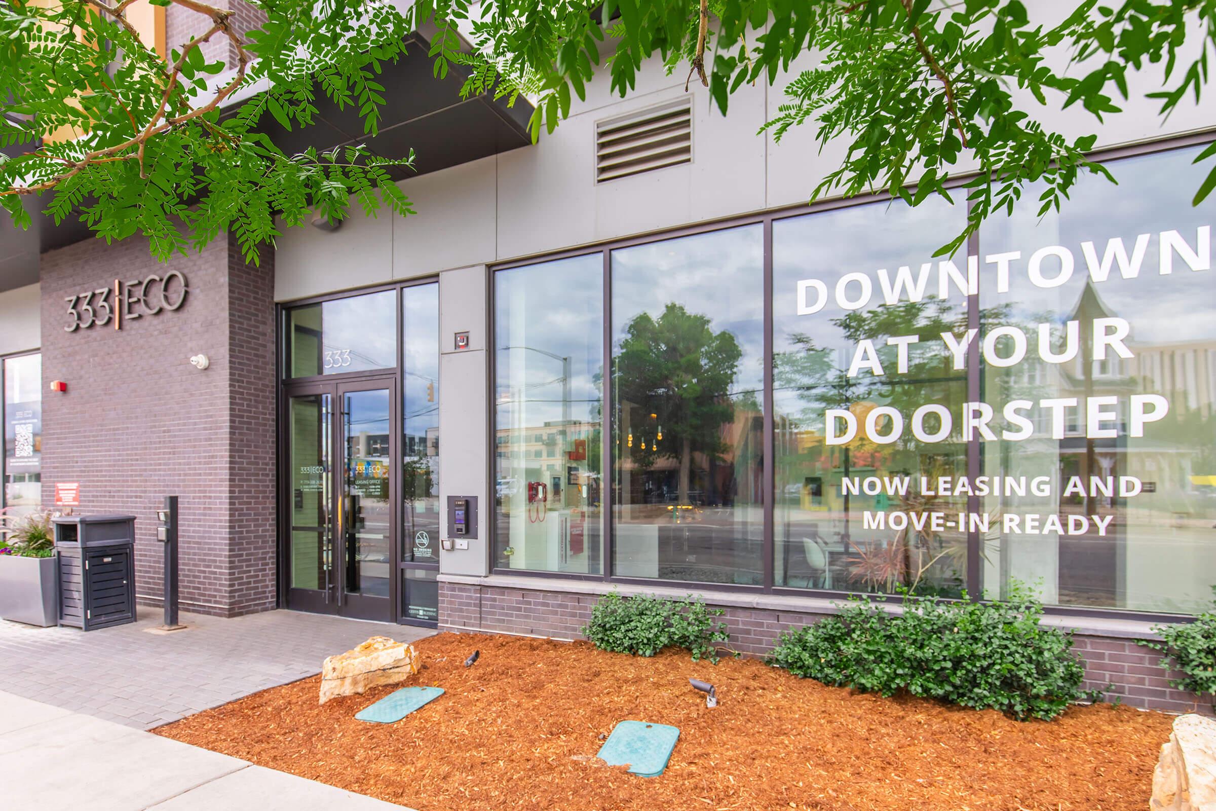 A modern building entrance with large glass windows displaying the text "Downtown at Your Doorstep. Now Leasing and Move-In Ready." The exterior features brickwork, landscaping with mulch and plants, and a mailbox mounted to the wall. The overall ambiance suggests a welcoming atmosphere for potential residents.