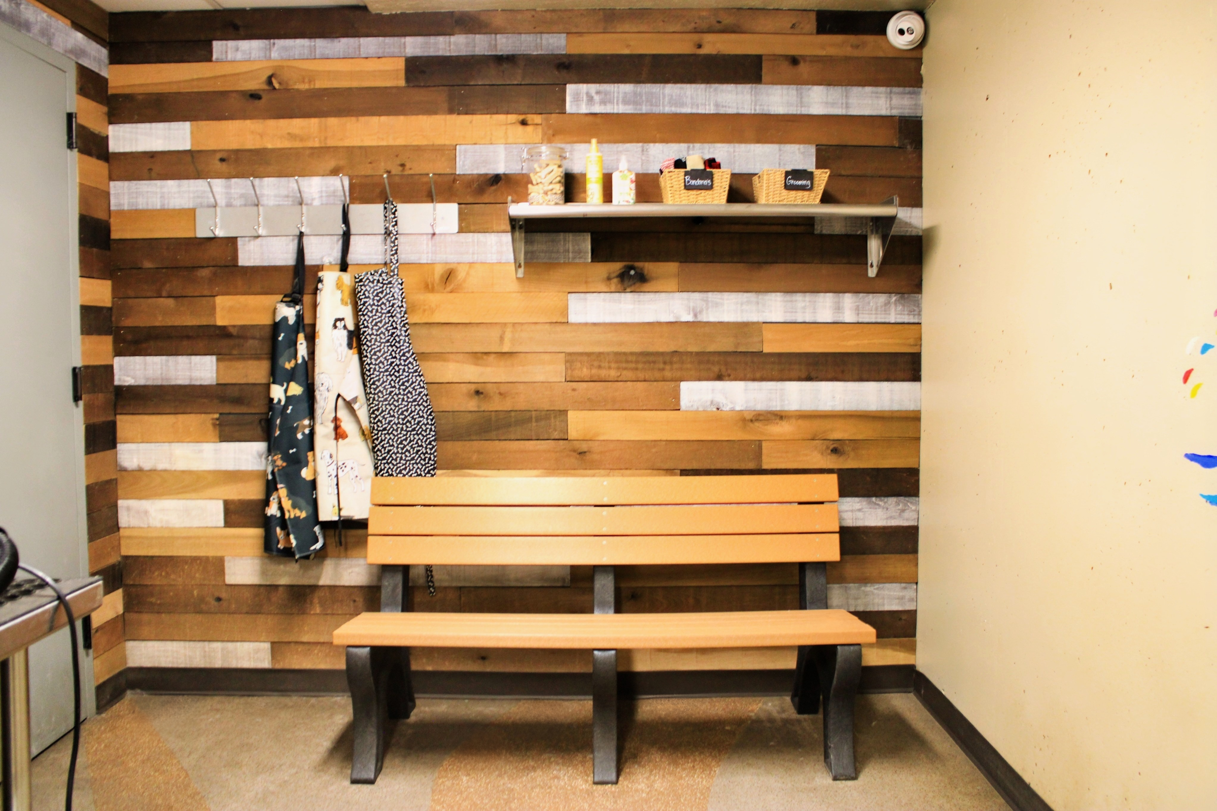 A wooden bench sits against a rustic wall made of various wooden planks. Above the bench, there are baskets on a shelf and several items hanging from hooks, including aprons. The setting has a warm, inviting atmosphere, suggesting a creative or communal space.