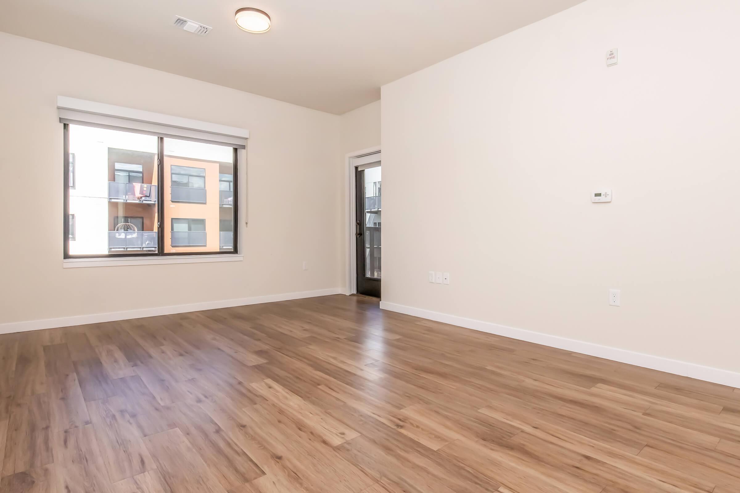 A spacious, empty room with light-colored walls and wooden flooring. There is a large window on one wall, showcasing views of nearby buildings. The room features a ceiling light and a door leading to a balcony or outdoor area. Ideal for a living space or office setup.