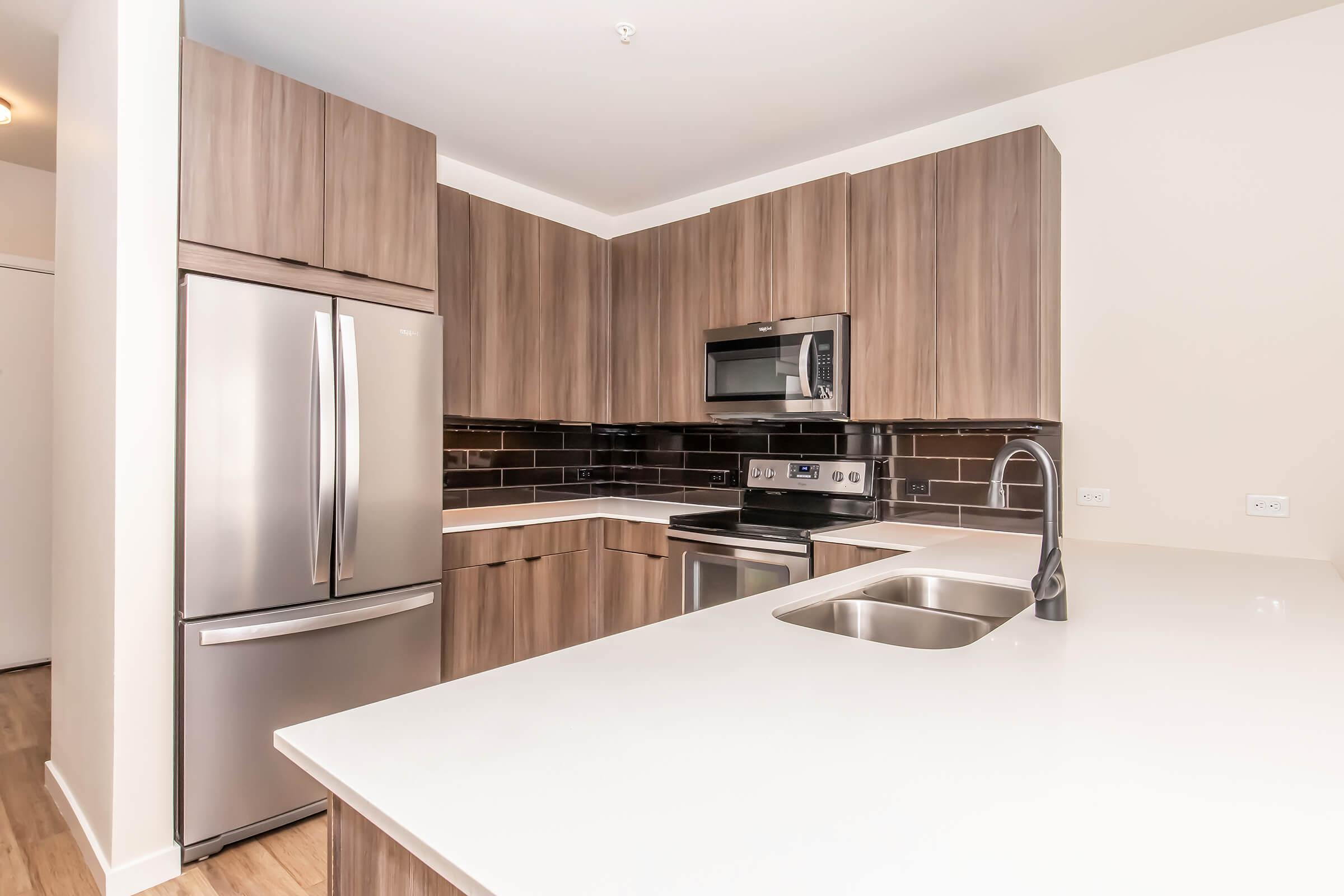 Modern kitchen featuring wooden cabinets, stainless steel appliances including a refrigerator, oven, and microwave. The countertop is white with a built-in sink. Dark backsplash tiles add contrast. The overall design is sleek and contemporary, with warm wood tones and a tidy layout.
