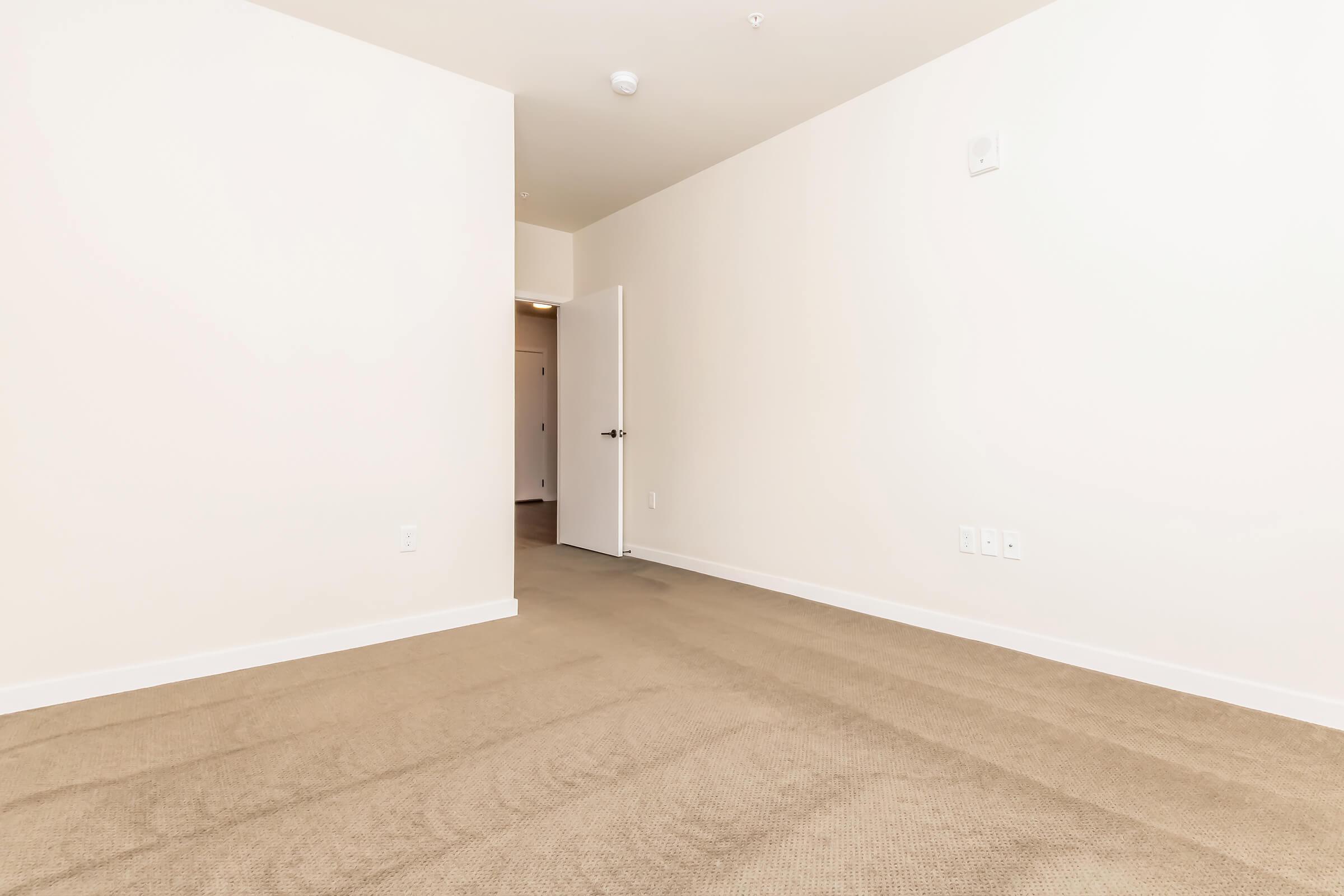 A spacious, empty room with beige carpet and light-colored walls. There is a white door on the right leading to another area, and the room is well-lit. The space is versatile and can be used for various purposes, such as an office or living area.