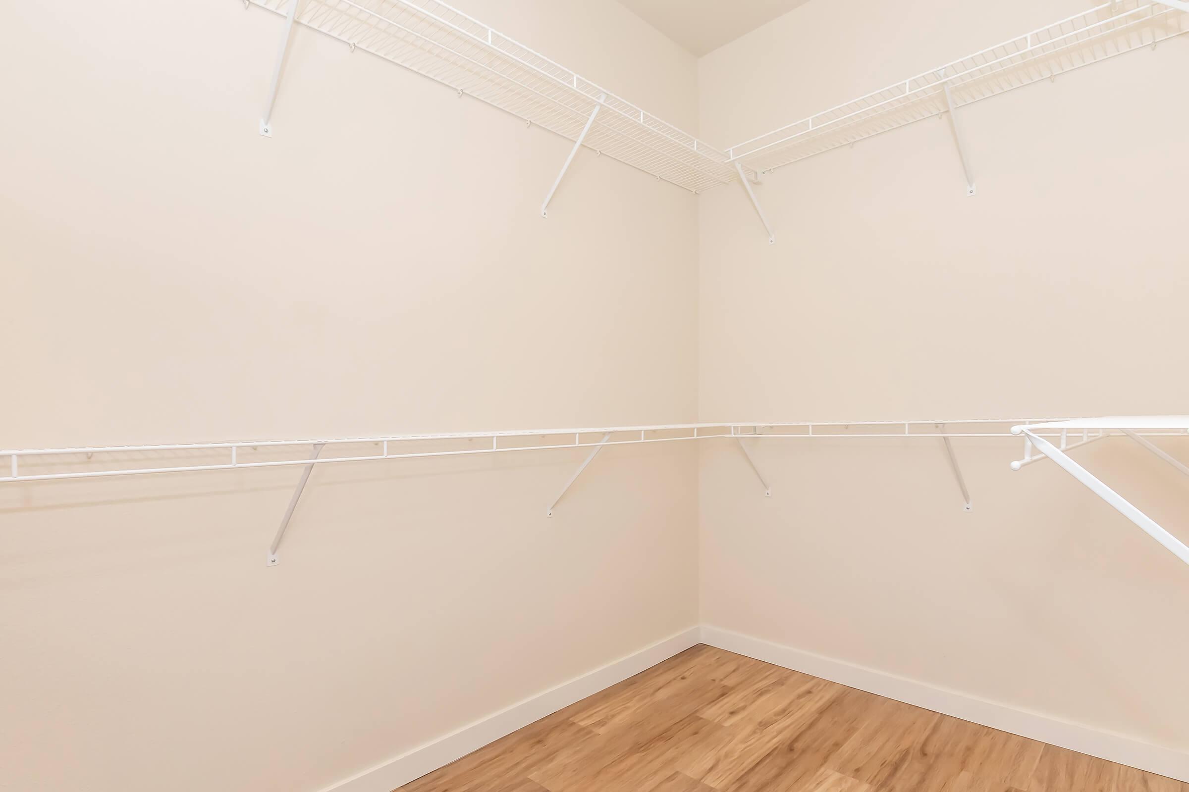 A spacious, empty closet featuring white wire shelving along two walls, with a light-colored wall and wooden flooring. The closet is organized with ample storage space available for clothes and accessories.