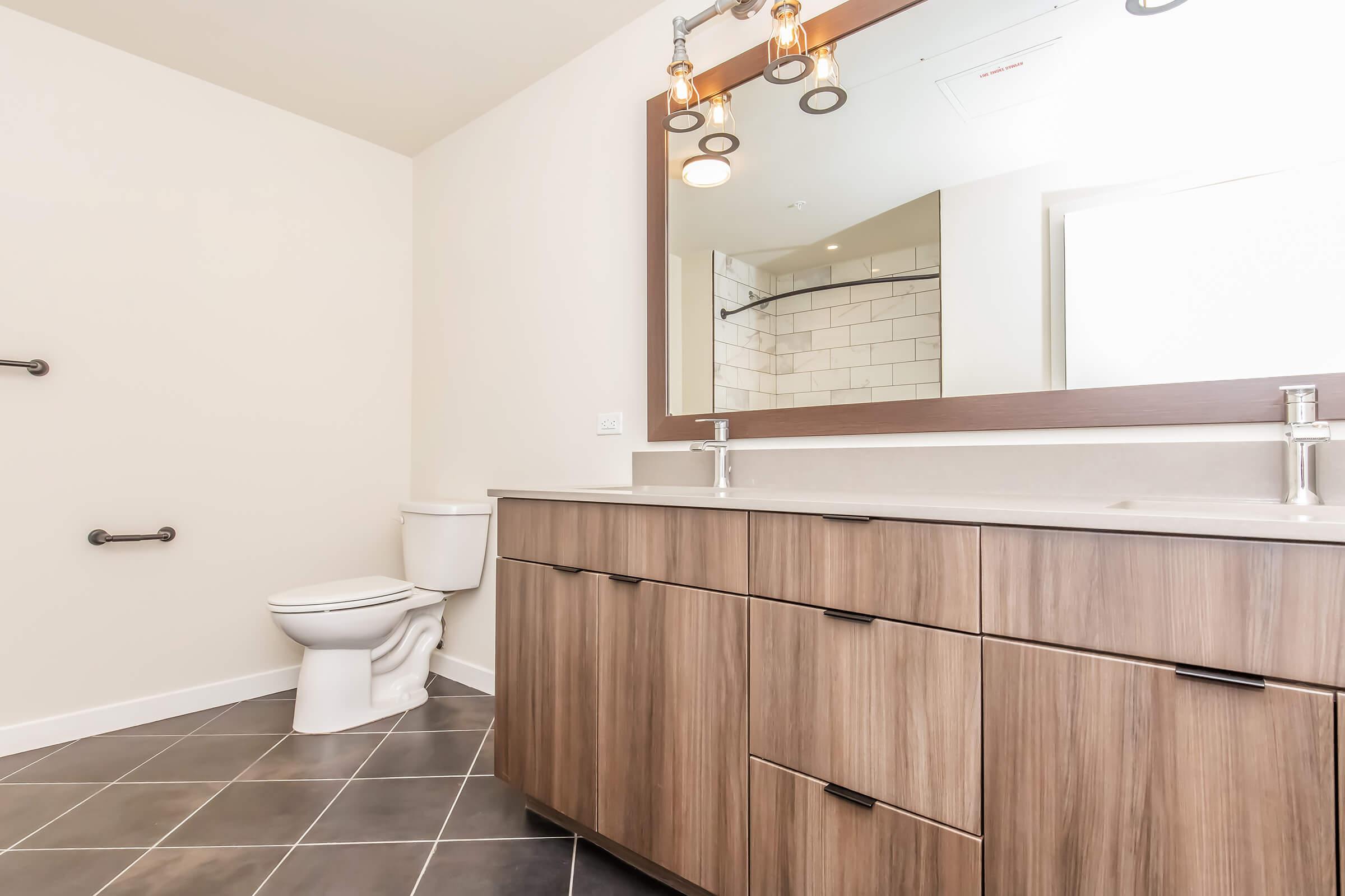 Modern bathroom featuring a large mirror above a stylish vanity with wooden cabinetry. A white toilet is positioned to the left, while sleek black tile flooring enhances the contemporary design. The space is well-lit with elegant light fixtures above the mirror, creating a warm and inviting atmosphere.
