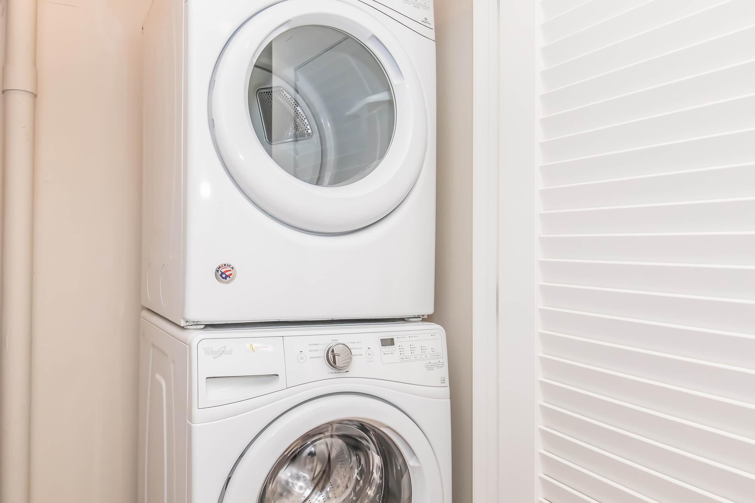 A stacked washer and dryer unit in a laundry space, featuring a white finish. The dryer, perched above the washing machine, has a glass door that allows visibility of the interior. The space is organized with light-colored walls and a linen closet beside the appliances.