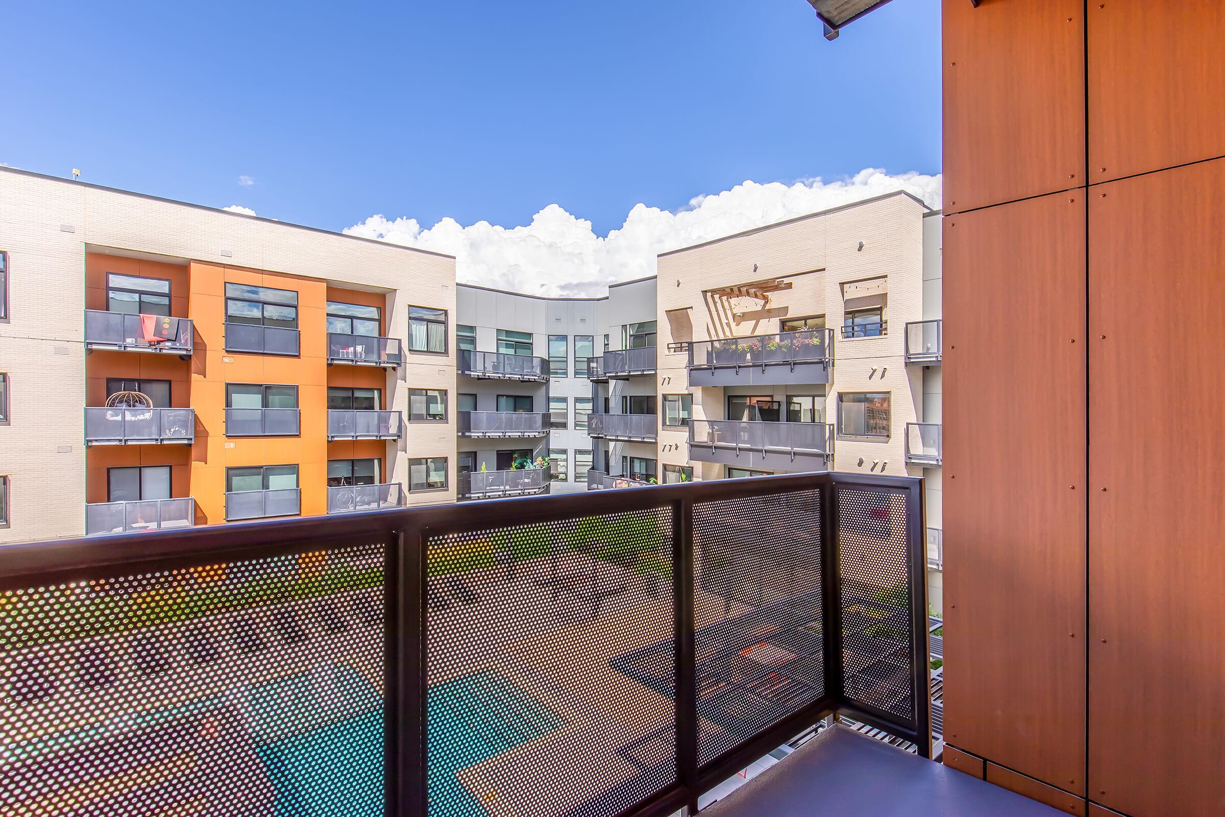 View from a balcony overlooking a modern apartment complex. The image features two multi-story buildings with a mix of beige and orange facades, balconies, and a swimming pool visible in the courtyard below. The sky is clear with fluffy clouds.