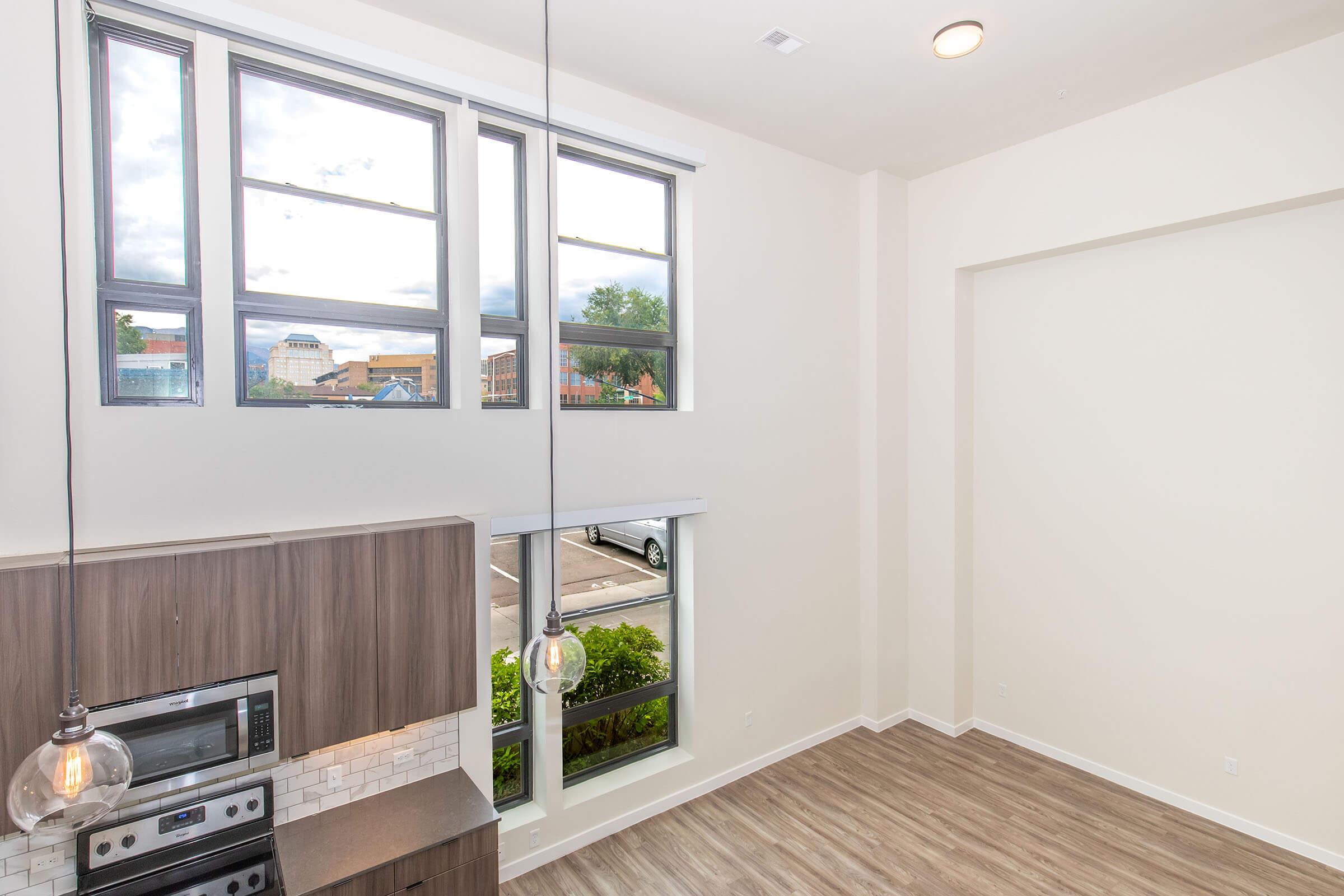 Bright, modern interior of a living space featuring large windows allowing natural light to flood in. A kitchen area with sleek cabinetry and stainless steel appliances is visible, and the flooring is a light wood finish. There’s a view of the outside with greenery and a street scene.