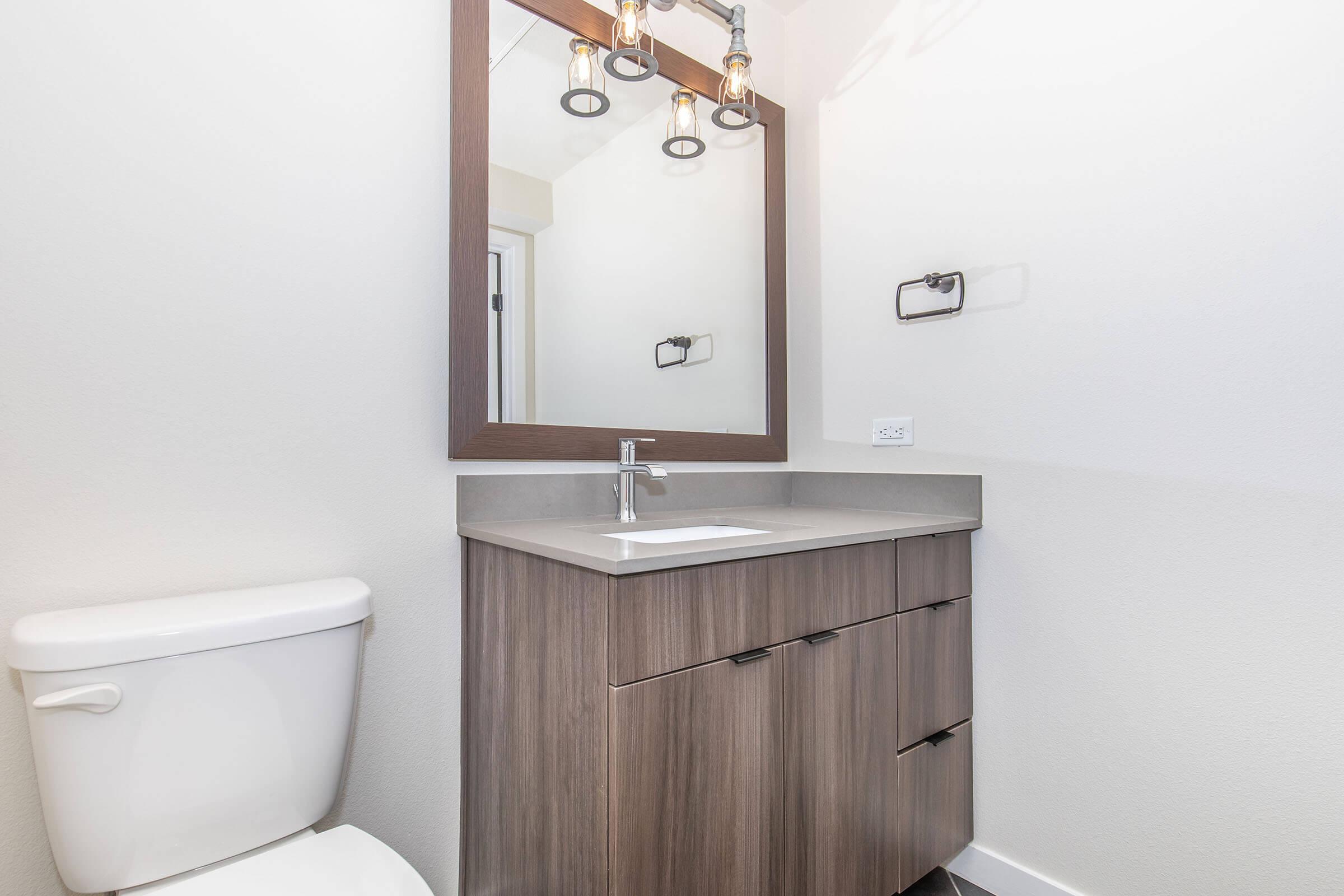 A modern bathroom featuring a compact vanity with a sink, wooden cabinets, a large mirror with decorative lighting, a toilet, and neutral-colored walls. The space is well-lit and designed for functionality and aesthetic appeal.
