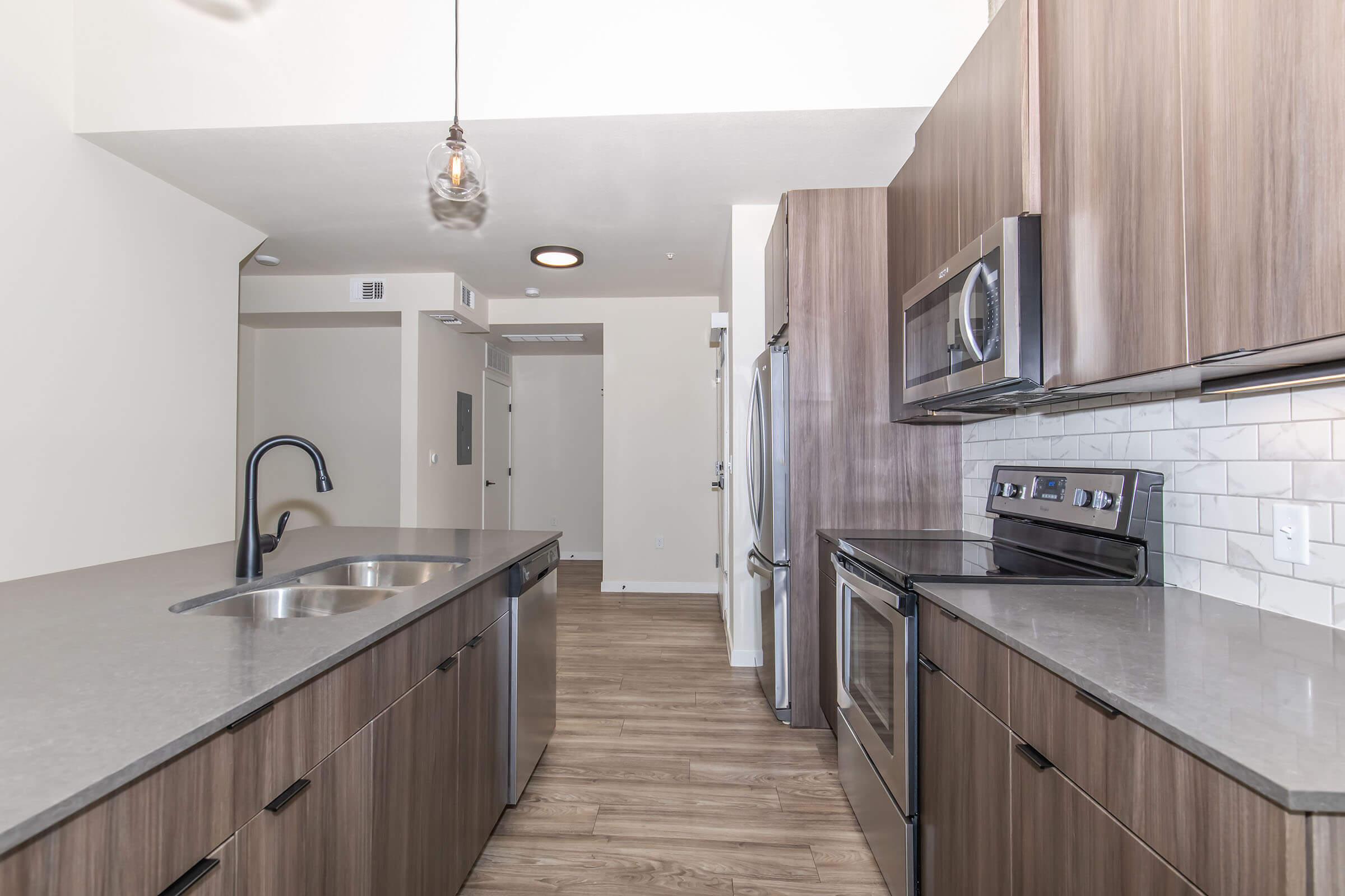 Modern kitchen featuring wooden cabinets, stainless steel appliances, and a large countertop with a double sink. The space includes a pendant light and an open layout leading to a bright area beyond. The flooring is a warm wood finish, complementing the contemporary design.