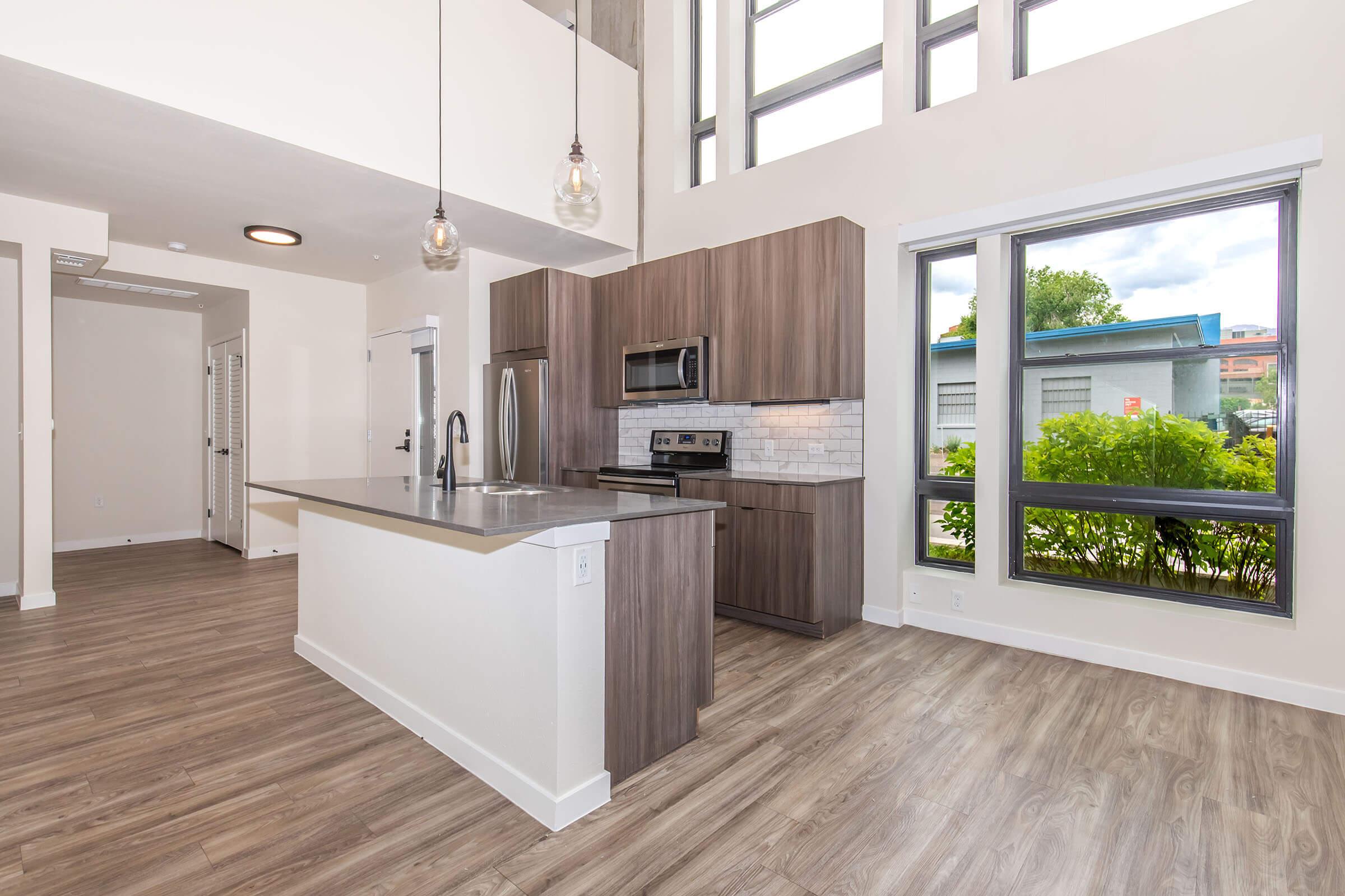 Modern kitchen in an open-concept living space featuring wooden cabinetry, stainless steel appliances, and a spacious island with seating. Large windows allow natural light to fill the room, showcasing a view of greenery outside. The flooring is a light wood finish, creating a warm and inviting atmosphere.