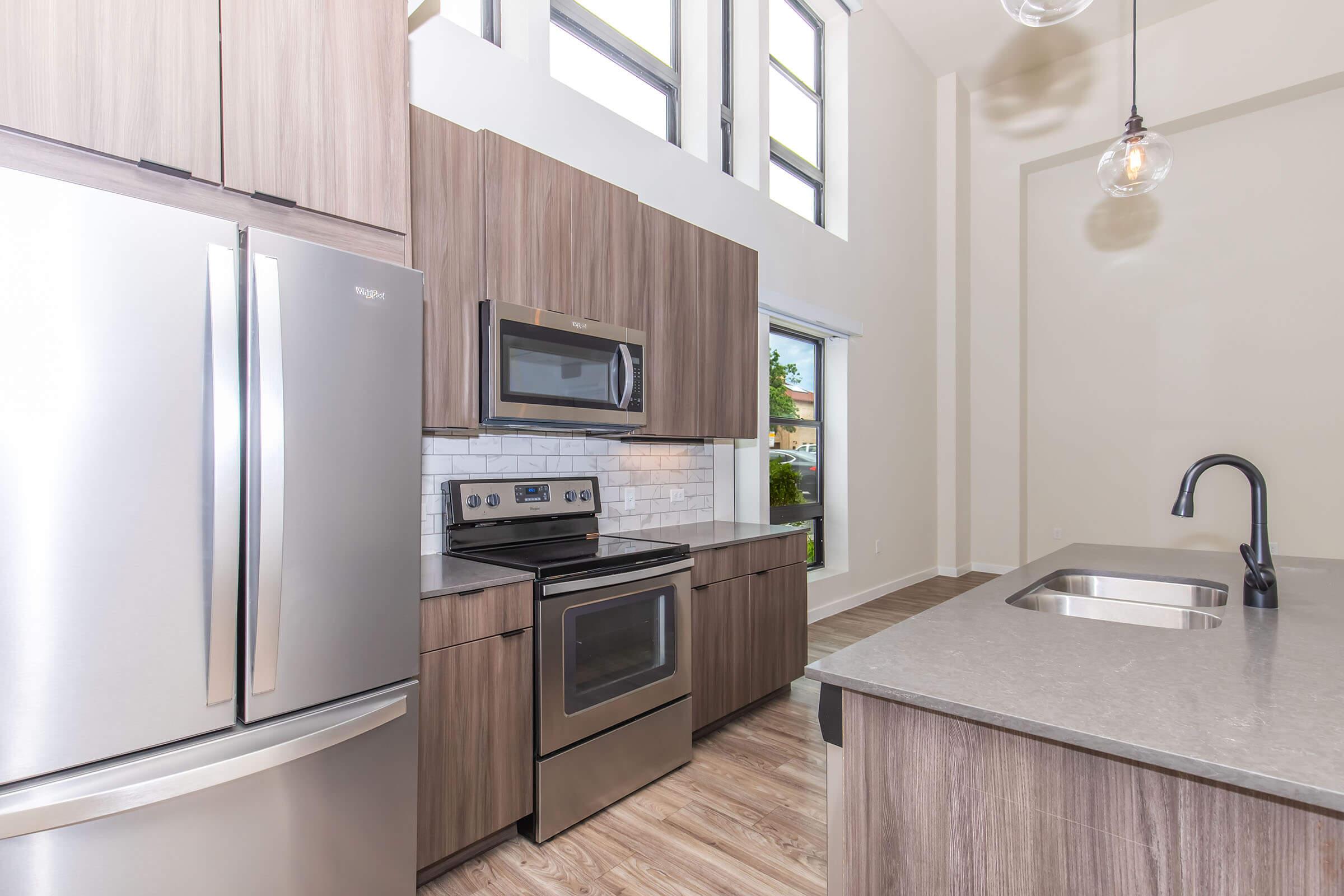 Modern kitchen featuring stainless steel appliances including a refrigerator, oven, and microwave. The cabinetry is in a warm wood finish with a gray countertop. Large windows let in natural light, and the space has a contemporary open layout with a sleek sink and pendant lighting.