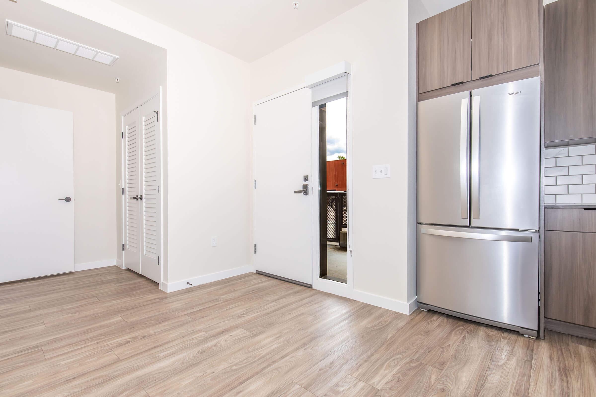 Interior view of a modern kitchen area featuring a stainless steel refrigerator, a door leading outside, and light wood flooring. The walls are painted in a neutral tone, and there are cabinets with a wood finish. A frosted glass door is visible on the left, enhancing the contemporary design.