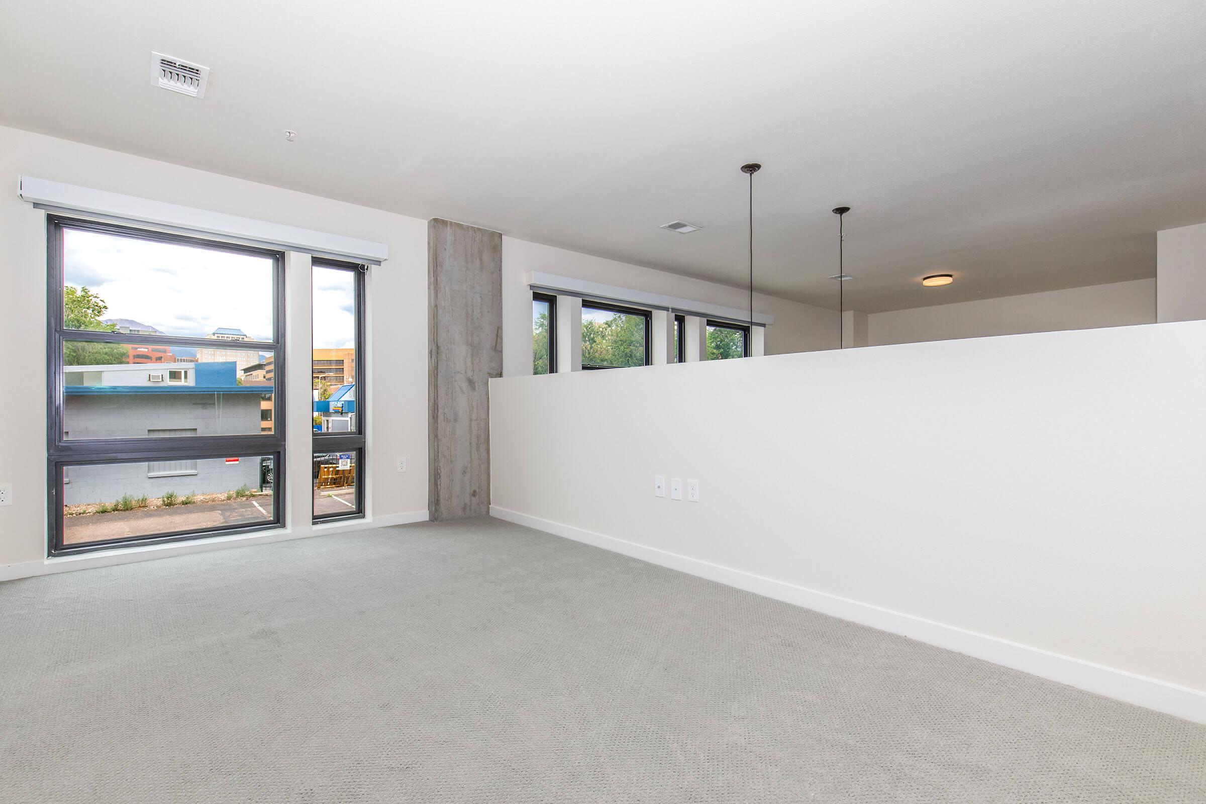 A spacious, empty room with light gray carpet and large windows letting in natural light. One side has a partial wall separating it from a kitchen area, while the ceiling features two pendant lights. The walls are painted in a soft, neutral color, creating a bright and open atmosphere.