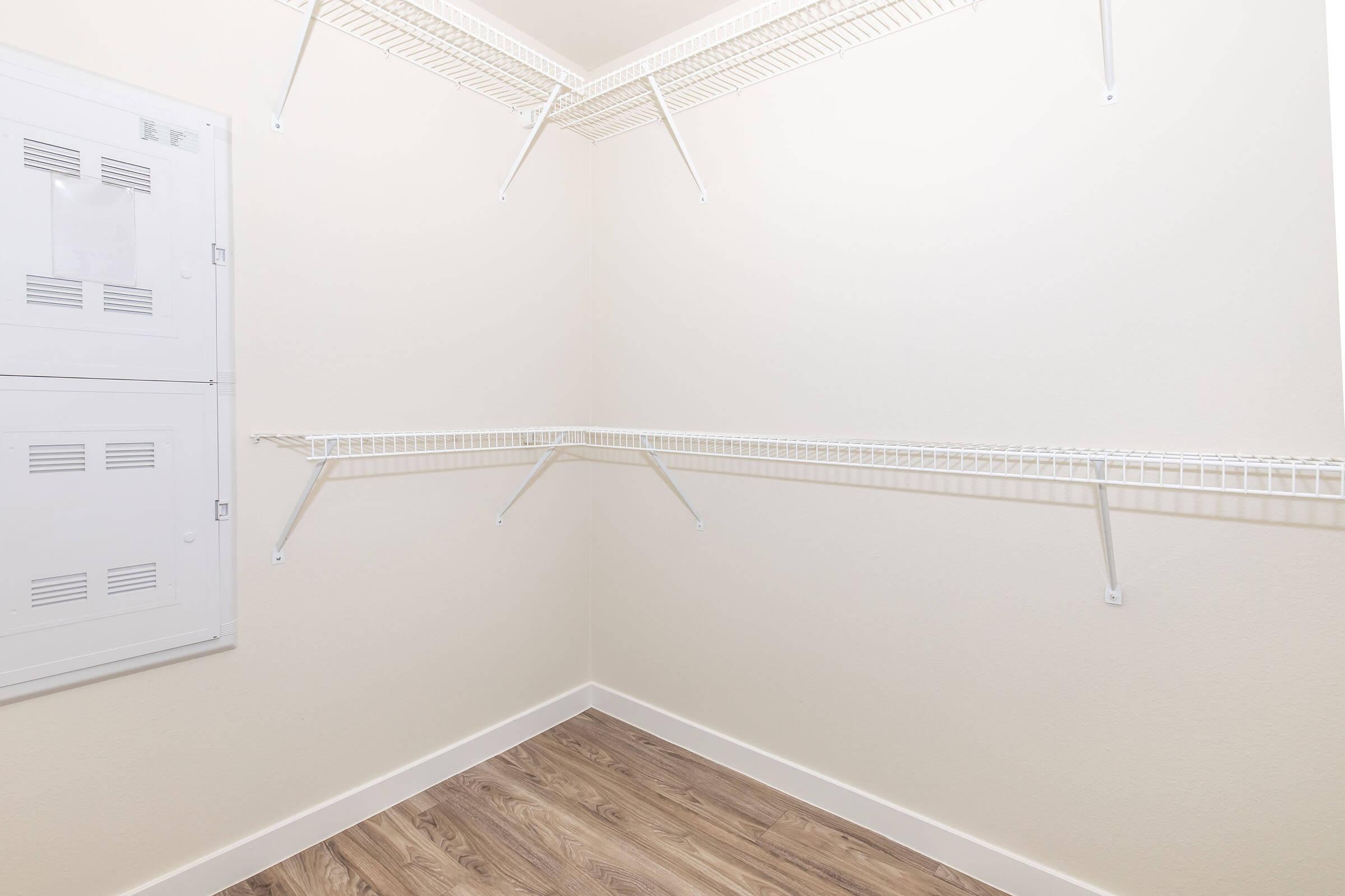 Empty closet with white wire shelving arranged in a corner. Walls are a light beige color, and the floor features light wood laminate. A utility panel is visible on the left side of the image. The space appears bright and clean, with no items stored on the shelves.