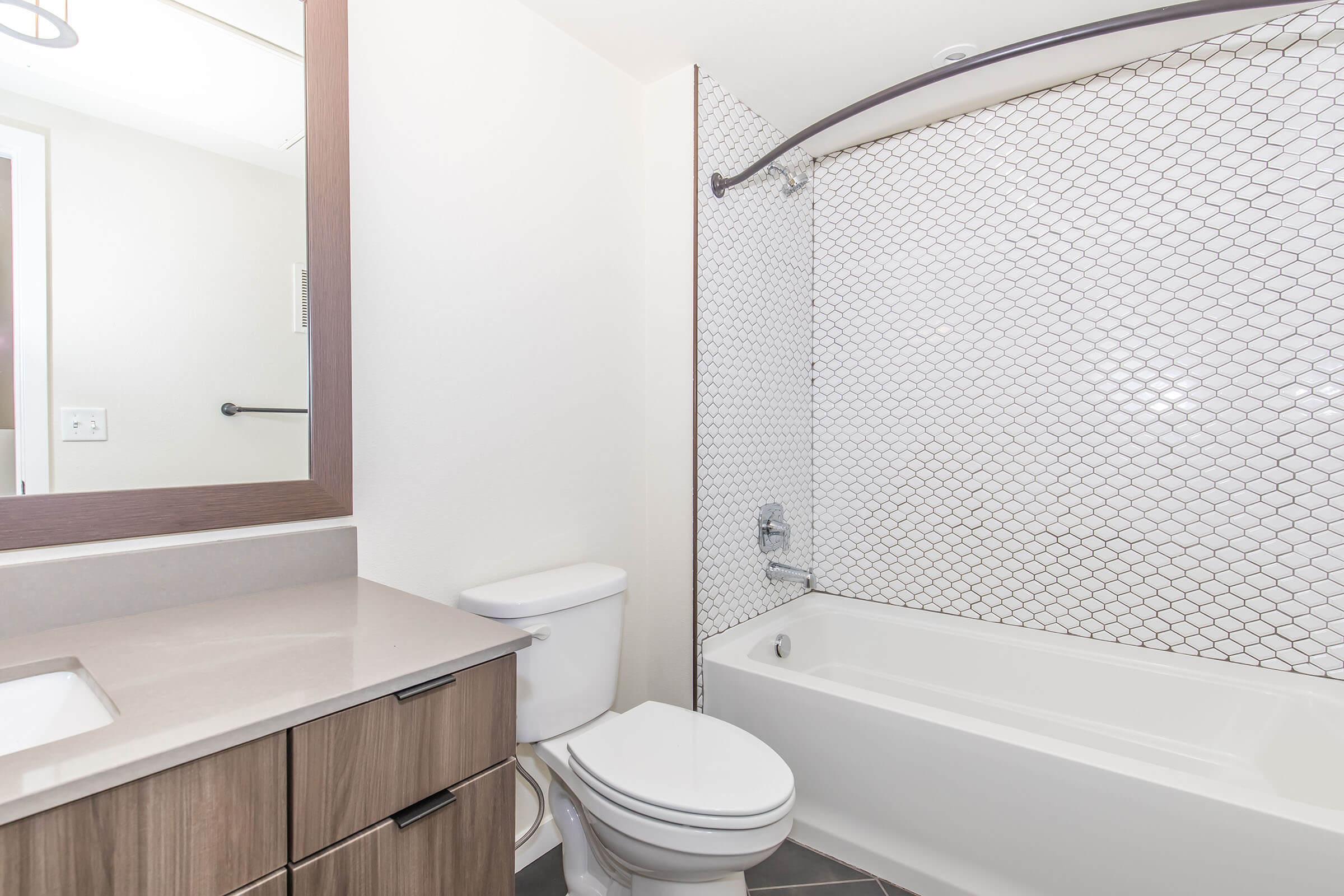 A modern bathroom featuring a bathtub with a shower, a tiled wall with a geometric pattern, a toilet, and a wooden vanity with a sink. A large mirror is mounted above the sink, and the space is illuminated by natural light. The overall design is clean and contemporary.