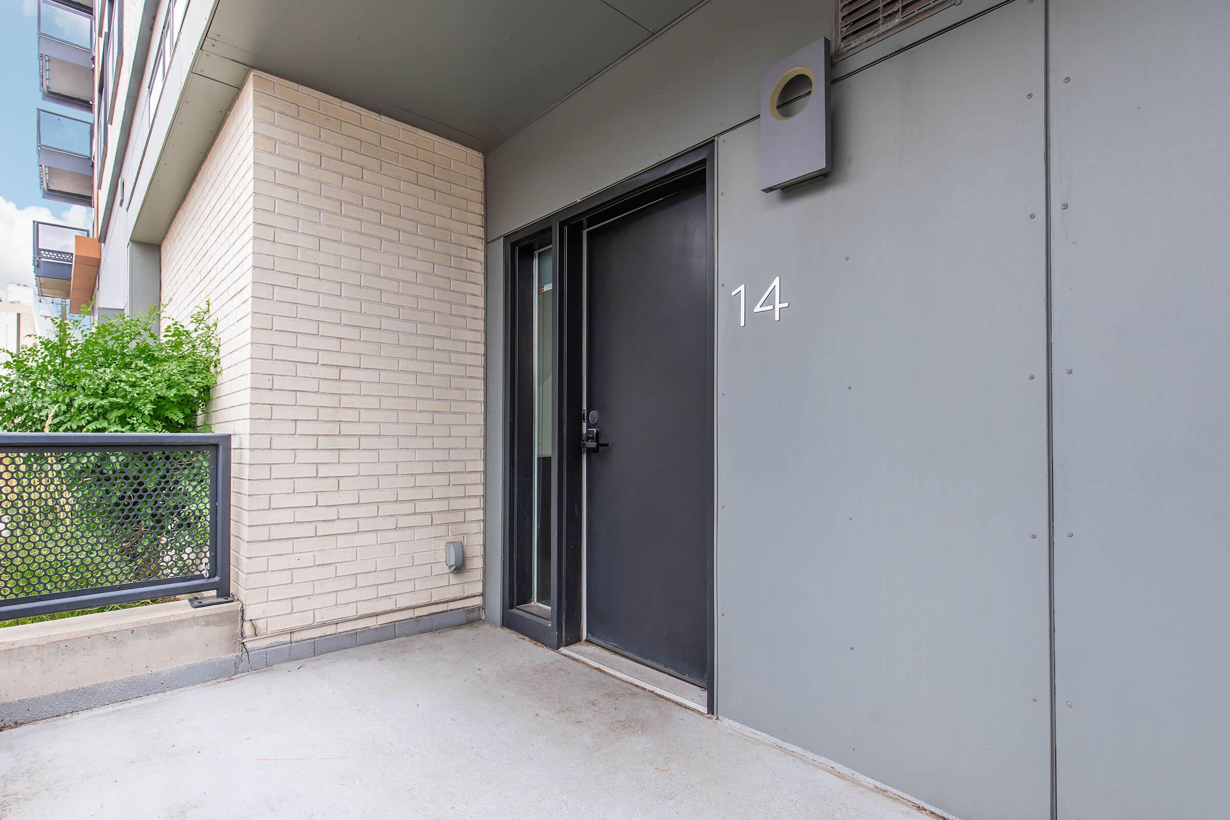 A modern building entrance with a gray exterior. The door is black with the number "14" displayed next to it. There's a small round light fixture mounted beside the door, and a small green plant visible in the background on the left. The area is clean and well-maintained.