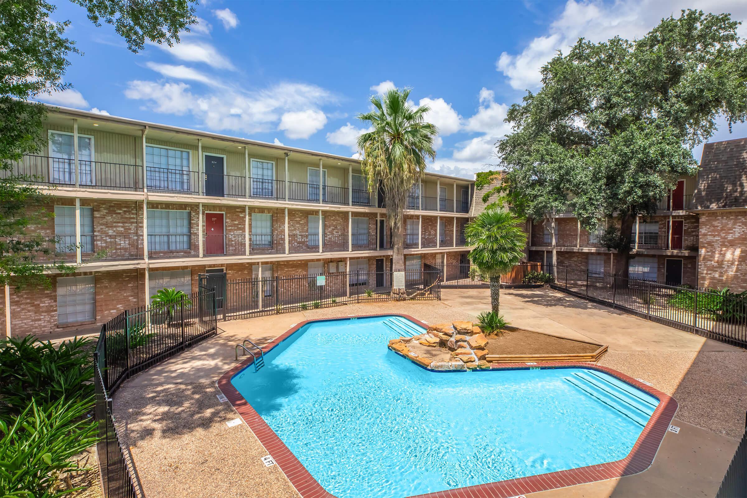 A sunny view of an apartment complex featuring a centrally located swimming pool surrounded by palm trees. The pool area has a rock formation and lounge chairs, with brick buildings and balconies visible in the background under a blue sky with scattered clouds.