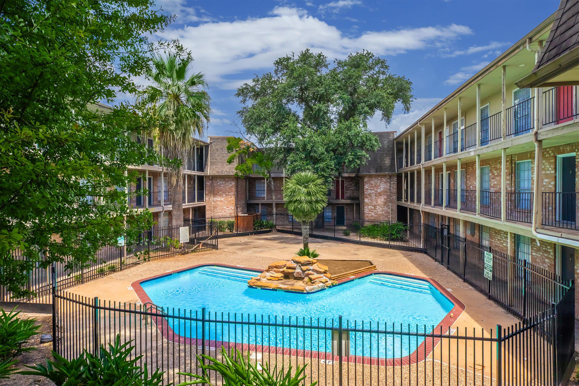 A clear blue swimming pool surrounded by a fenced area, with palm trees and a large oak tree in the center. The pool is bordered by a small rock formation, and there are two-story buildings with balconies on either side, set against a bright blue sky with a few clouds.